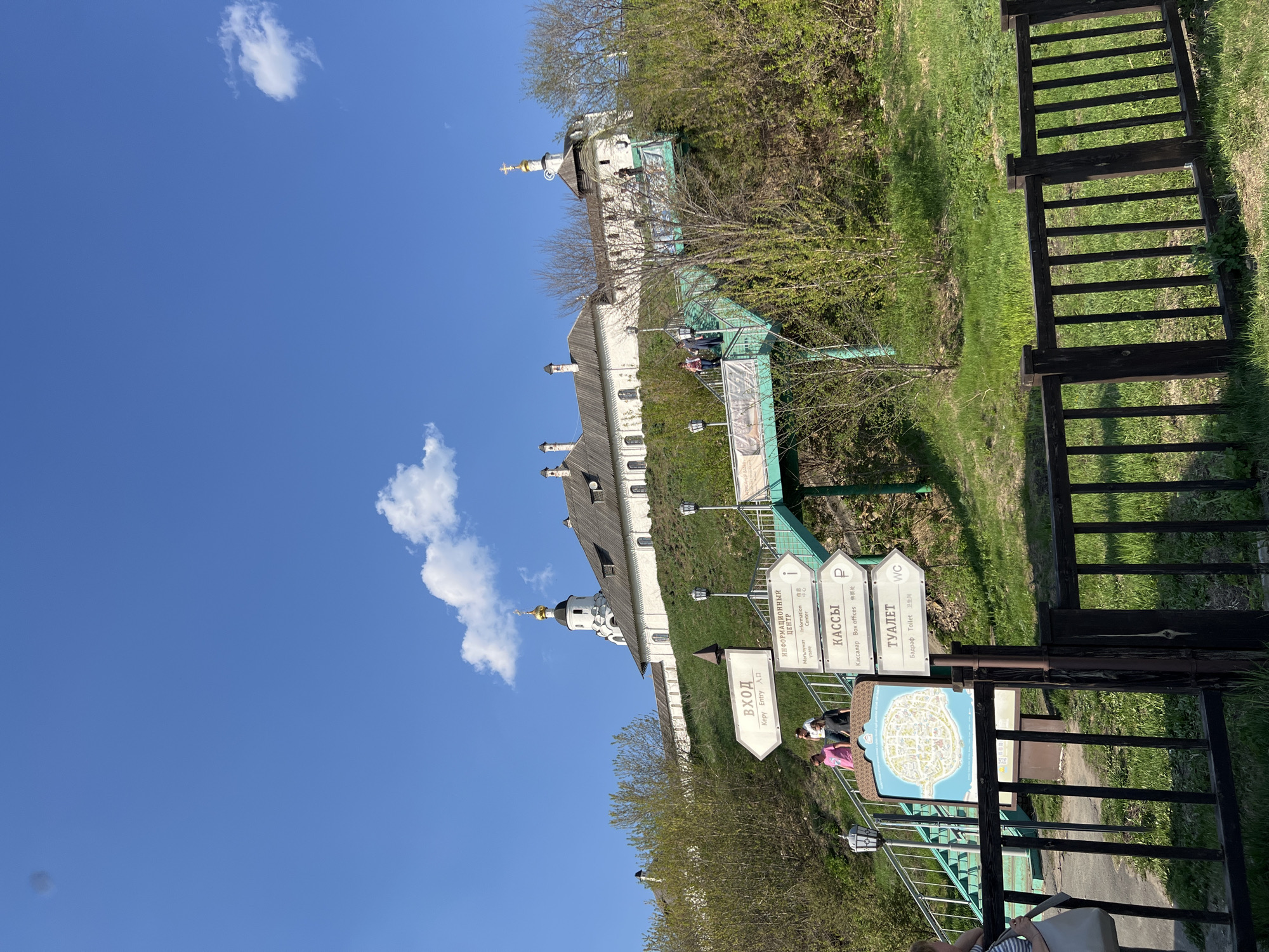 A daytime, outdoor, ground-level shot displays a Russian Orthodox Church complex in Sviyazhsk, Russia.
The white buildings with green roofs, situated along a slope, include multiple onion-shaped domes topped with gold crosses. A green walkway descends the hill toward the viewer, marked by signs in Russian and English, indicating "Entry," "Information Center," "Box Offices," and "Toilet". A map of the area is also visible. People are walking along the walkway.
To the right, a dark wooden fence runs along the walkway's edge, separating it from the grassy hillside. The clear blue sky contains scattered white clouds.