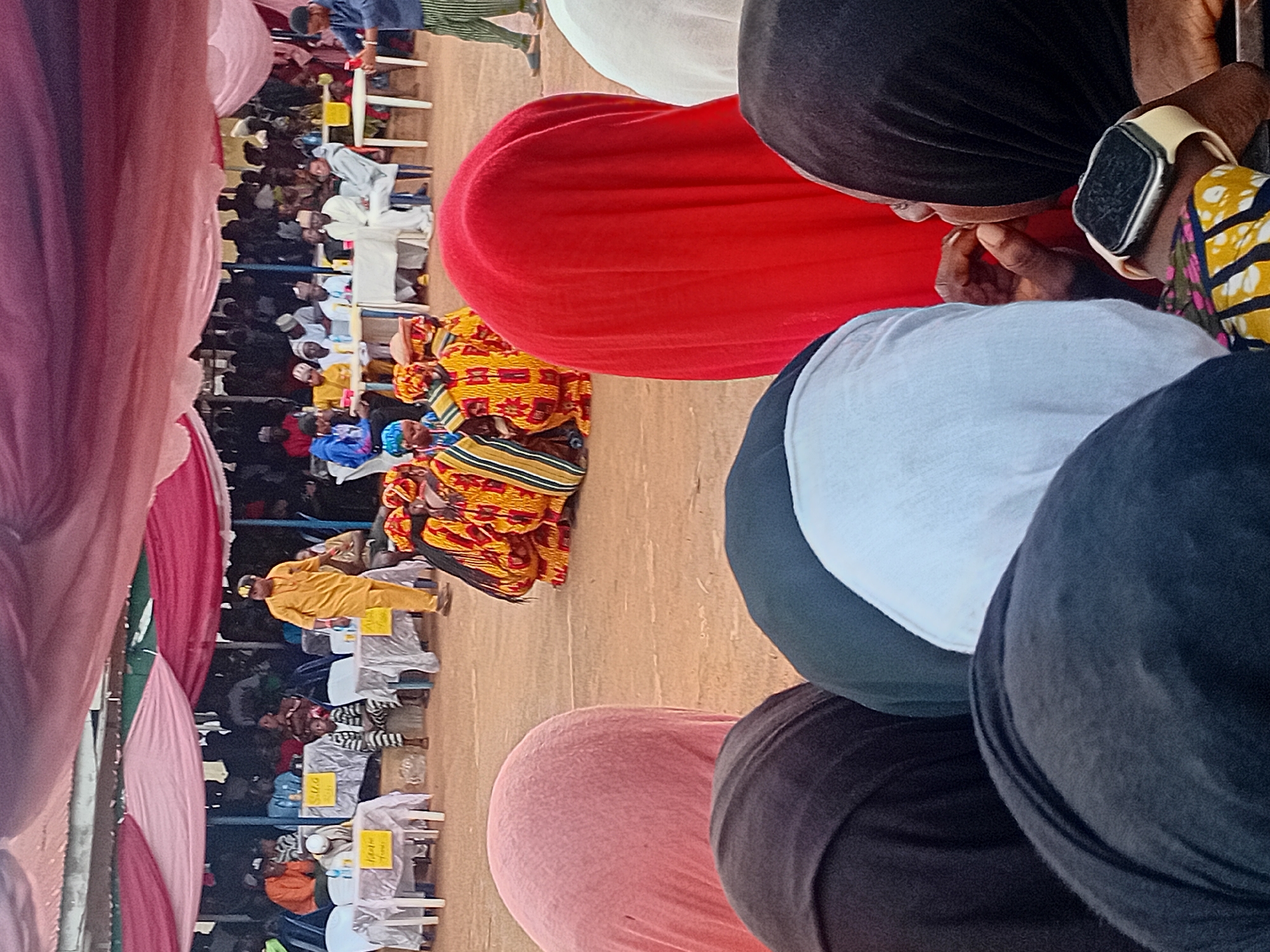 This image depicts an outdoor community event taking place in Ogbogbo, Nigeria. The scene is set under a temporary canopy structure, characterized by draped sections of light pink and dark red fabric overhead. The ground is unpaved, appearing as light brown dirt.
In the immediate foreground, several individuals are visible from behind, facing away from the camera, indicating they are spectators. From left to right, their backs show varied attire: a dark top, a person wearing a pink head covering, another dark top, a person in a dark green top, a white top, a person in a vibrant red head covering, and a person in a white head covering. On the far right, an individual wearing a black head covering is partially visible from the side, with their right hand near their mouth. This person wears a multi-colored patterned sleeve (yellow, black, red, blue) on their left arm, and an Apple Watch with a light-colored band is visible on their left wrist.
In the midground, a group of approximately five to six individuals are crouched or kneeling on the dirt ground. They are dressed in elaborate, matching traditional or ceremonial attire, predominantly yellow and red with distinct striped patterns. This group appears to be performing or engaging in a structured activity. To the right of this group, a man in a blue long-sleeved shirt and green-and-white striped pants stands, observing the activity. Further to the left, a man in a full yellow traditional garment walks across the midground, with his back partially turned to the viewer.
The background reveals an audience area where numerous people are seated at white plastic tables covered with white tablecloths. Small plastic bottles, likely containing water, are visible on these tables. Some tables bear yellow signs with text; partially legible examples include "S.U.O." and "Alum." The attendees are dressed in a mix of traditional and casual clothing. The canopy structure is supported by visible poles, and the overall environment suggests an organized public gathering.