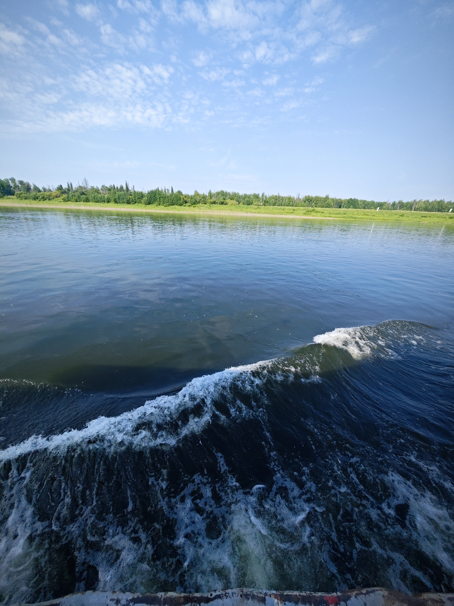 A wide shot captures a body of water, possibly a river or lake, with a distinct wake in the foreground indicating movement. The water is a deep blue-green, reflecting the sky above. A foamy white wave crashes on the right side of the frame, trailing into turbulent water. In the distance, a lush green shoreline is visible, lined with trees and vegetation under a bright blue sky dotted with scattered white clouds. The scene suggests a calm day, possibly during summer, with the perspective implying the photo was taken from a moving vessel. There are no people or animals visible in the image. The specific location is the city of Novonazimovo, Russia.