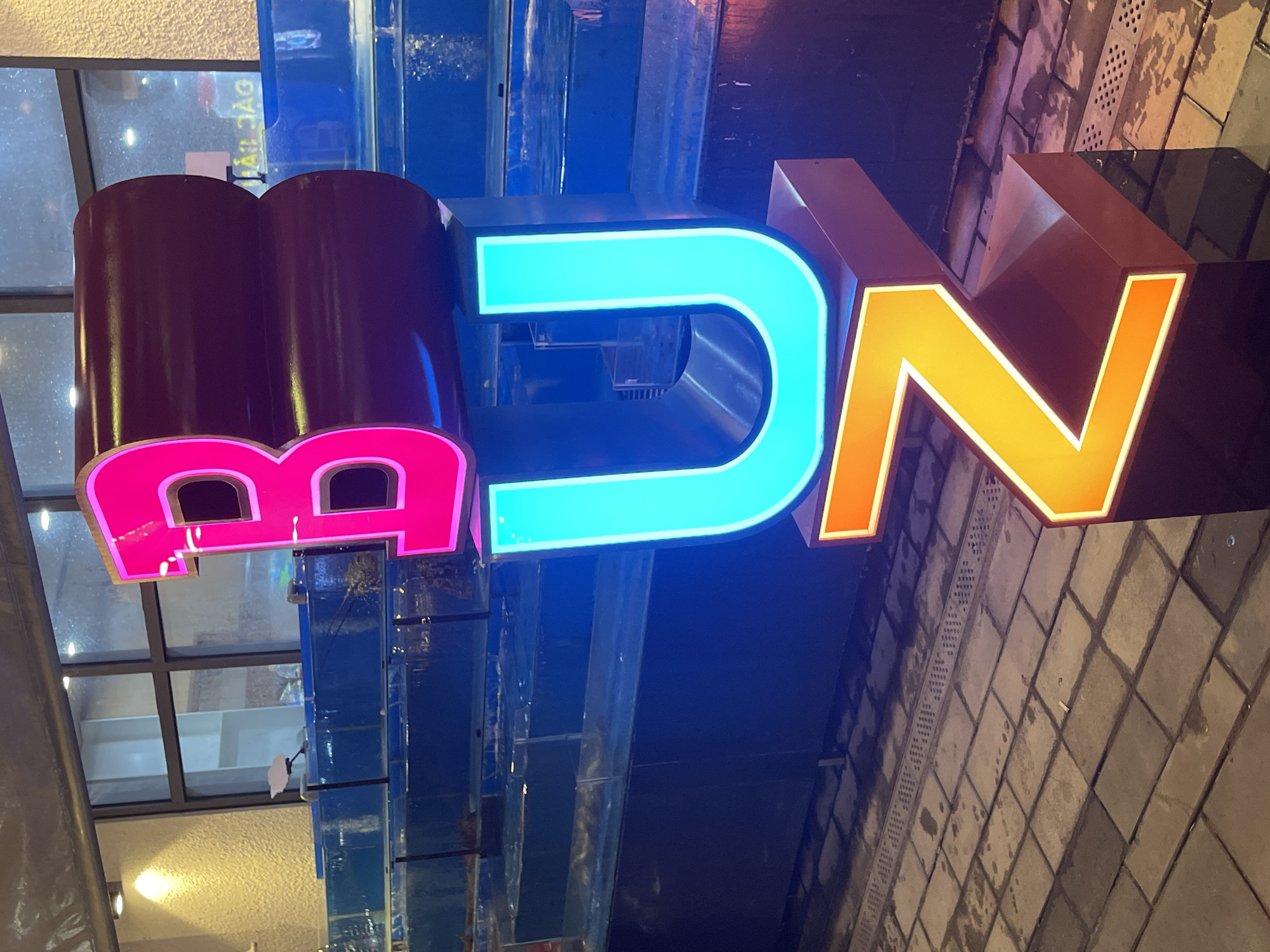 The image depicts a brightly lit, three-dimensional sign spelling out "BUN" in large, stylized letters. The letter "B" is a vibrant pink, the "U" is a neon cyan, and the "N" is a glowing orange, all outlined in white. These letters are stacked vertically. The sign appears to be placed outdoors, in front of a building with large windows and a textured, cream-colored wall. Behind the sign, rows of illuminated blue tanks are visible, suggesting a seafood restaurant or market. The ground is paved with rectangular stone tiles, wet and reflecting the lights. In the background, through the windows, a sign with Vietnamese text, "HẢI SẢN," which translates to "seafood," is discernible, confirming the location as a seafood establishment. The overall scene suggests a commercial establishment at night in Đà Nẵng, Vietnam, likely a restaurant that specializes in seafood, indicated by the tanks and the signage. There are no people visible in the image.