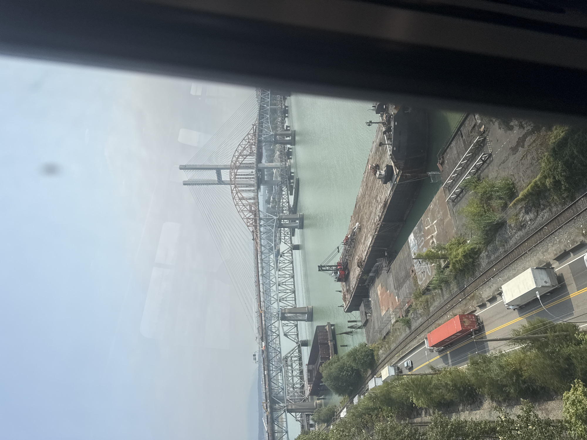 The image captures a wide view of the Fraser River in New Westminster, Canada, featuring multiple bridges and industrial activity.
In the foreground, a large barge with a crane is docked at a pier. Adjacent to the pier are railway tracks and a road where several trucks are visible. One truck is bright red, and another is white. Trees line the left side of the road and tracks.
Spanning the river are two distinct bridges. The closer bridge is a large, multi-span steel truss bridge. Further in the distance, a modern cable-stayed bridge with prominent towers and diagonal cables dominates the skyline. The river itself is a greenish hue.
The sky is bright and hazy, suggesting either a warm day or smoke from wildfires. The overall scene depicts a busy port and transportation hub with significant infrastructure. No people are discernible in the image.