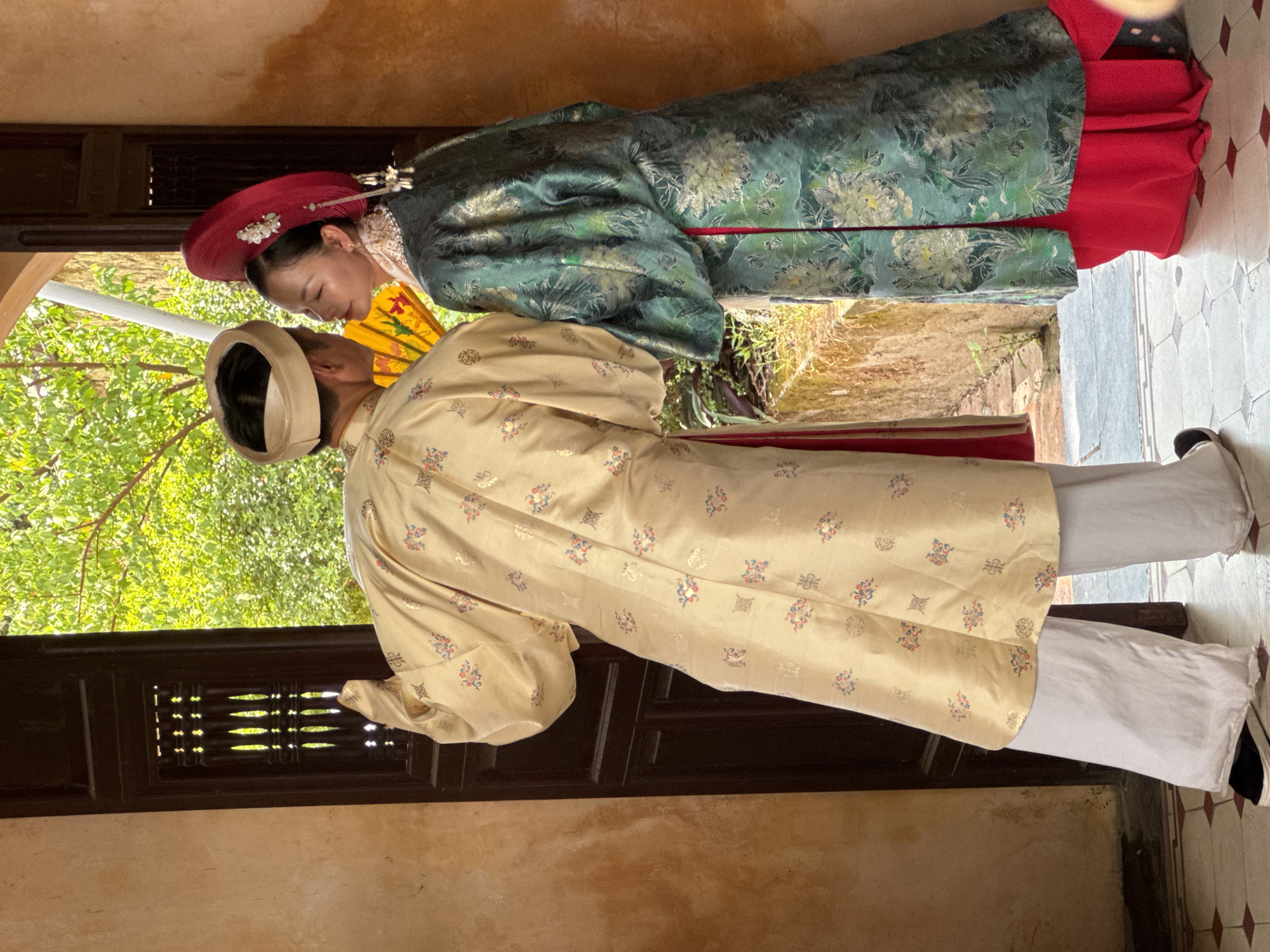 Two individuals, likely a couple, are positioned in a doorway, facing each other and leaning in as if for a kiss. The person on the left, dressed in a light yellow, ornate robe and white trousers, turns their back to the viewer and is wearing a head wrap. The person on the right, facing the viewer, wears a vibrant green, patterned robe over a red skirt and a large, circular red hat adorned with white flowers. They hold a yellow fan. The doorway is dark wood, with latticework on the left panel. Beyond the doorway, lush green foliage is visible under bright daylight. The floor inside is tiled with a diamond pattern, and the wall to the left is textured and a warm ochre color. The setting appears to be historical or culturally significant, possibly within a traditional Vietnamese dwelling or courtyard in Phú Xuân Ward, Vietnam.