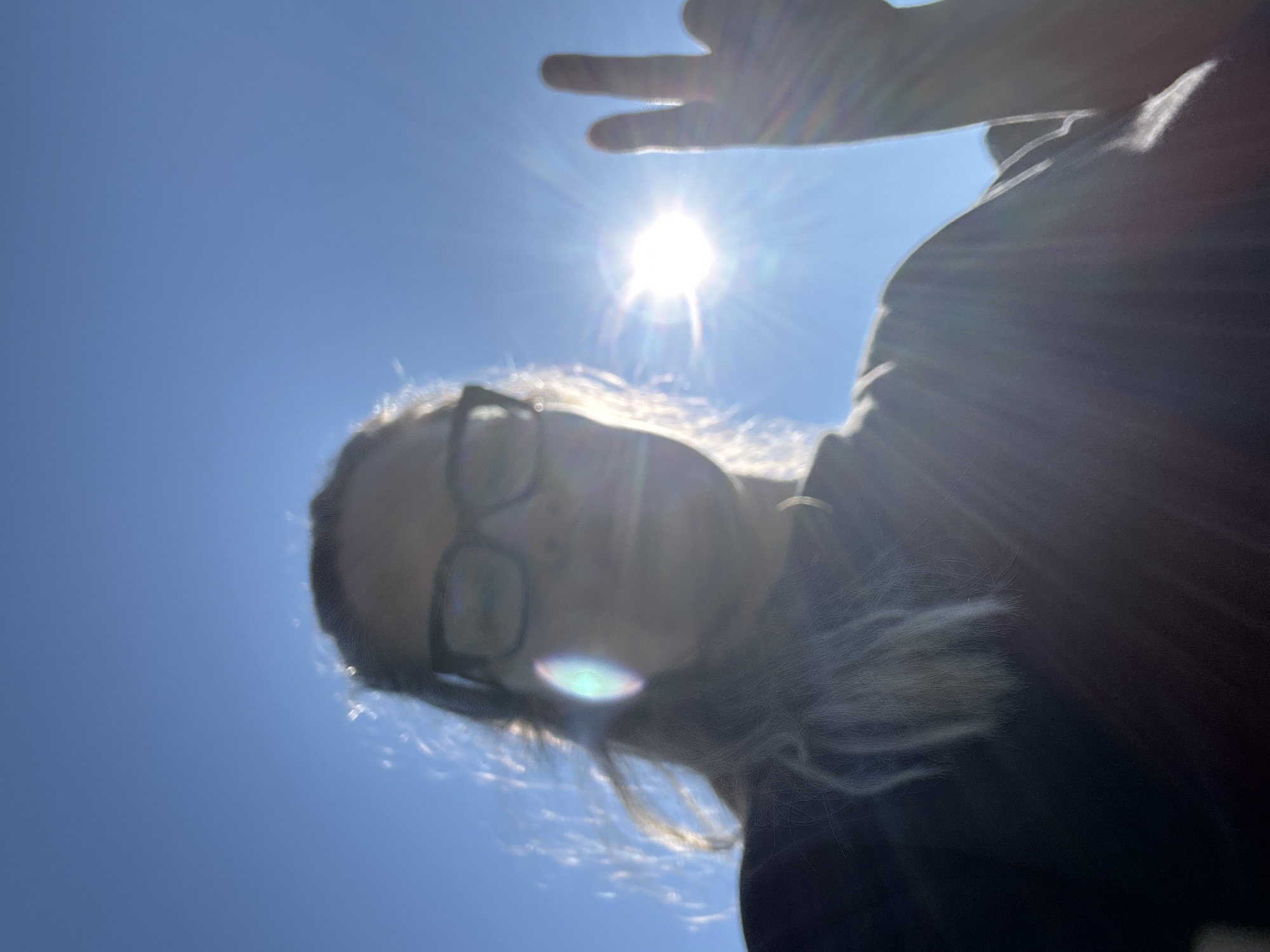 In this low-angle, backlit selfie taken outdoors against a bright blue sky, a person wearing glasses and a dark shirt is partially visible. Their face is obscured by the intense sunlight, but their hair, pulled back, is also visible. The person is making a peace sign with their right hand, with two fingers extended. The sun is prominently featured, emitting strong lens flares that create a starburst effect. The environment appears to be an open space with clear skies. The location is not specified within the image itself, and therefore the requested location of Калицино, Russia, cannot be confirmed from the visual evidence.
