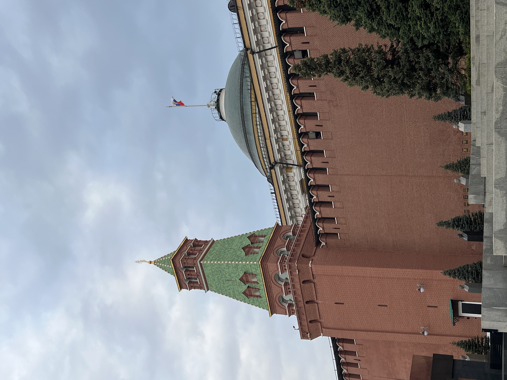 The image shows a view of the Moscow Kremlin in Russia. Specifically, it features a red brick building with a green-roofed tower and a white and gold dome structure. The Russian flag is visible atop the dome. The sky is overcast. Parts of the surrounding architecture and landscaping are also present, including trees and steps.
