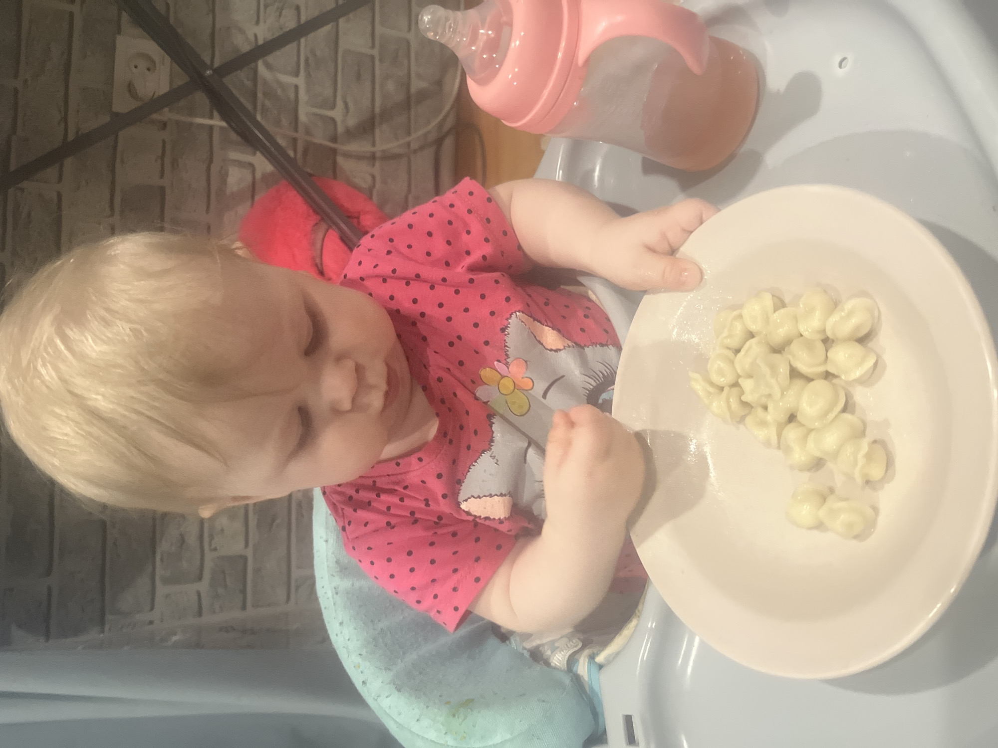 A top-down indoor shot shows a baby in a high chair with a plate of pasta and a bottle. The baby has blond hair and is wearing a pink shirt with black polka dots and a gray elephant graphic. The plate in front of the baby contains a pile of small, white pasta shapes. A pink baby bottle rests on the high chair tray. The high chair appears to be blue. In the background, there's a brick-patterned wall and a metal chair frame. The location is unknown and not specified in the image.