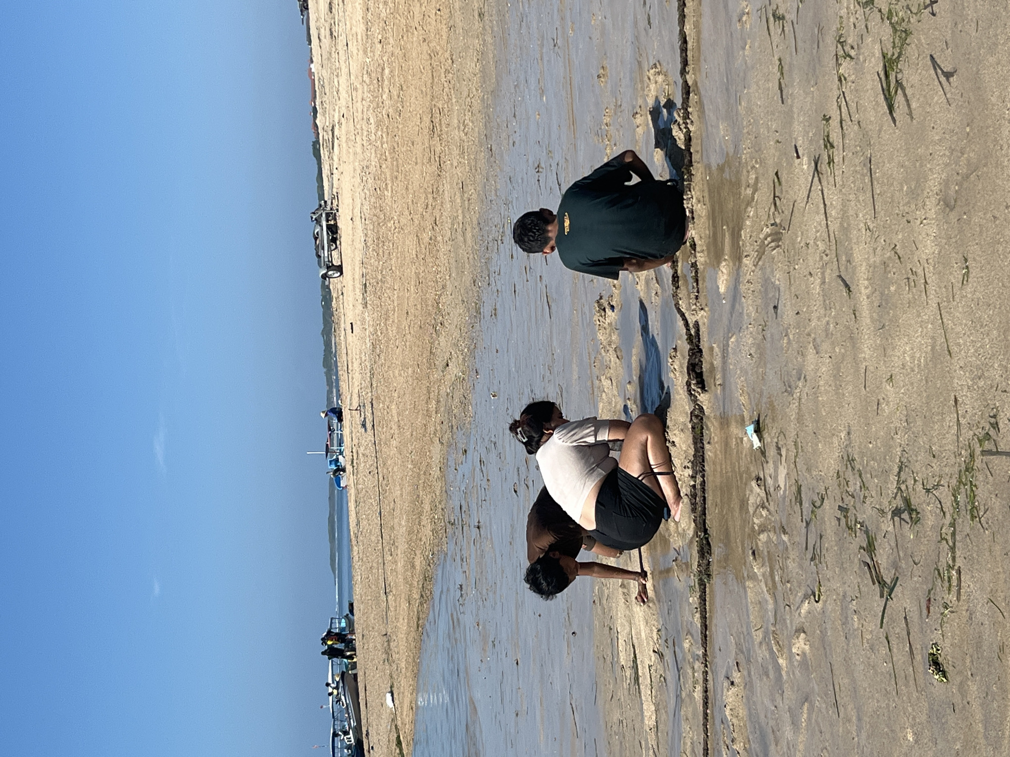 Three individuals are crouched on a sandy beach in Kuta Selatan, Indonesia. The person on the left, wearing a brown t-shirt, is leaning forward, appearing to interact with the wet sand. The person in the center, wearing a light-colored shirt and black shorts, is also looking down at the sand. The person on the right, wearing a dark green t-shirt, is sitting with their back to the camera. The beach is wet in the foreground with visible seaweed and water. In the background, there are several boats on the sand, some with people gathered around them, and a trailer. The sky is clear and blue.