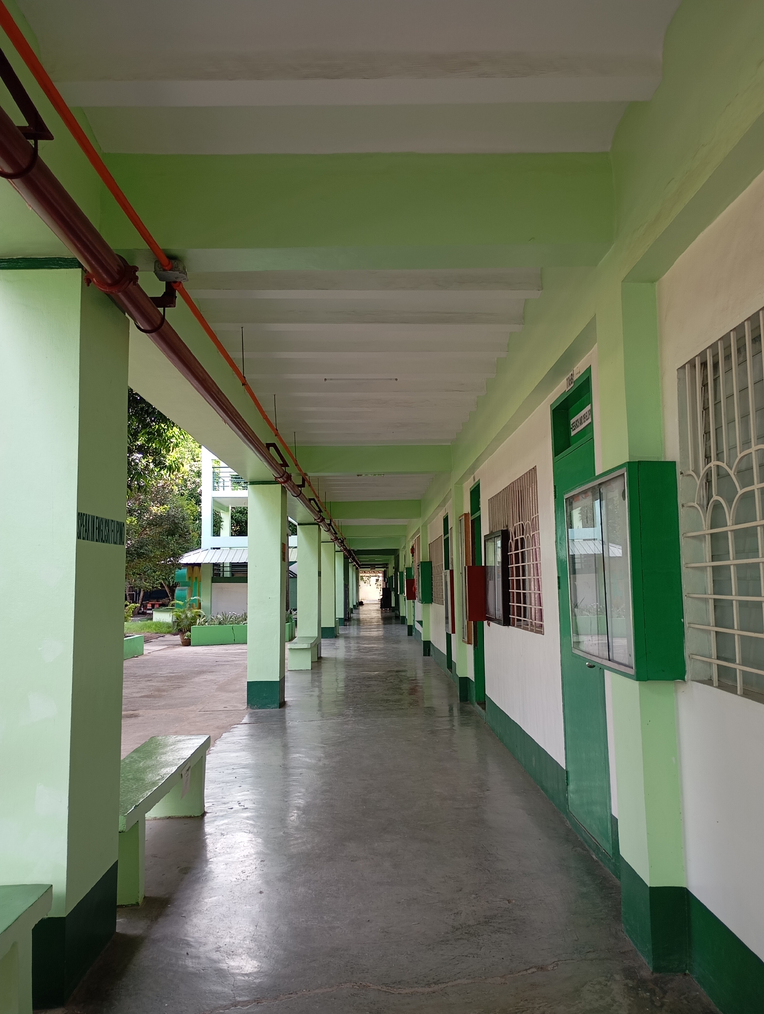The image captures a long, brightly lit outdoor hallway or corridor, characteristic of a school or institutional building, likely located in Davao City, Philippines. No people are visible, lending a quiet and orderly atmosphere to the scene.
The hallway features a consistent color palette of light green walls and pillars, accented with a darker green trim along their bases. The ceiling is white, traversed by visible utility pipes—one red and one orange—and white concrete beams. The floor is made of smooth, gray concrete, which reflects the ample natural light.
On the left side, light green concrete benches are placed alongside the pillars. Beyond these pillars, lush green trees and foliage are visible, suggesting an outdoor garden or courtyard, with a multi-story building further in the background. On the right side, a white wall with a dark green base contains several doors, windows with white security grilles, and green display cases or bulletin boards. One green door prominently displays the text "RESEARCH AND DEVELOPMENT" and the number "116B" above it. A partially visible vertical text, "SPEARHEAD", is also seen on a pillar on the left.
The presence of distinct shadows cast by the pillars and benches on the floor indicates a sunny daytime. The overall impression is that of a clean, well-maintained, and functional public or academic space.