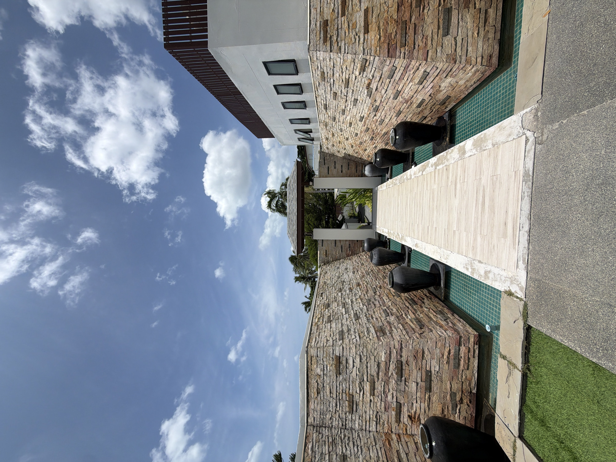 The image depicts an outdoor walkway in Ko Samui District, Thailand, leading towards a covered archway. The walkway is made of light-colored wooden planks and is flanked by a shallow pool with turquoise tiled bottom. Large black ceramic vases are placed at intervals within the pool. The walkway and pool are bordered by textured stone walls. To the right, a white building with a slatted wooden roofline features a row of dark-framed windows. Lush green grass is visible in the foreground on the left. The sky above is blue with scattered white clouds. The scene appears to be a tranquil and well-maintained outdoor space, possibly part of a resort or private residence.