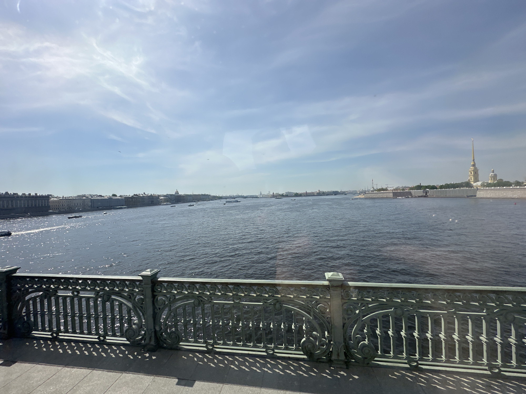 The image captures a view in Saint Petersburg, Russia, specifically along the Neva River, with the Peter and Paul Fortress visible on the right. The photo is taken from a bridge, as evidenced by the decorative green wrought-iron railing in the foreground. The river is wide, with several boats visible along its surface. On the opposite bank, a row of buildings lines the riverfront. The sky is mostly clear with scattered clouds. The perspective suggests the photographer is standing on the bridge's pedestrian walkway. Shadows cast by the railing are visible on the bridge's surface.