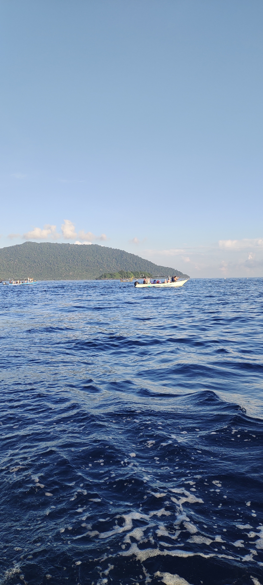 In the foreground of this image, the dark blue ocean exhibits rippling waves with white foam crests, indicating movement. Further into the scene, two boats traverse the water. The boat on the right is a white speed boat with a canopy, carrying several passengers. The boat on the left is a longer, blue vessel, also with multiple passengers. In the background, a large, verdant island rises from the sea, its slopes covered in dense vegetation. The sky above is a clear, pale blue, with a few wispy white clouds. The location is the waters off the city of Sabang, Indonesia, an archipelago known for its natural beauty and clear waters, often visited by tourists for island hopping and snorkeling.