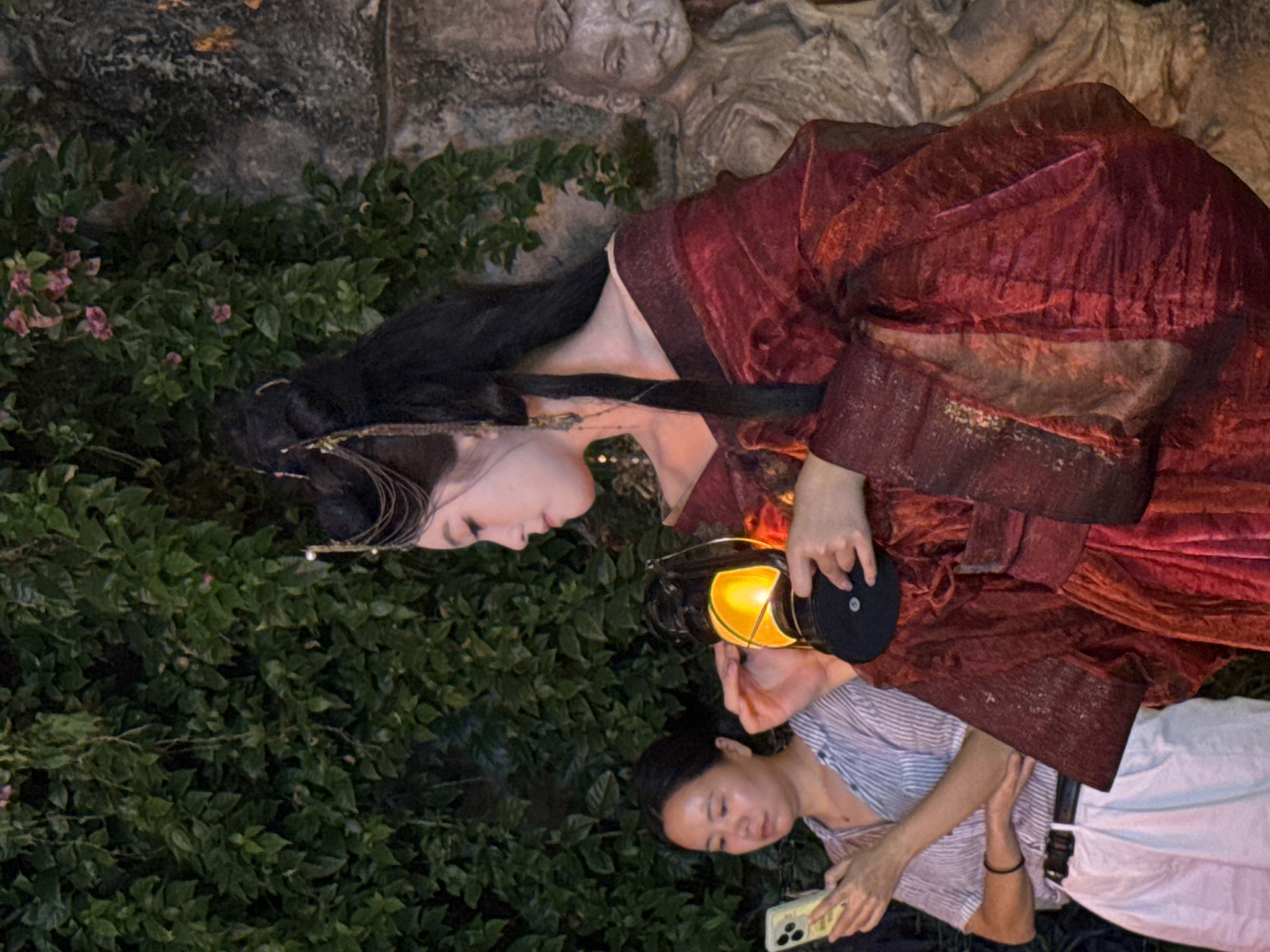 In the Yuzhong District of China, a young woman in traditional Chinese attire, a flowing red robe adorned with gold accents, holds an antique lantern emitting a warm glow. Her hair is intricately styled with ornate accessories, and she is looking down at the lantern. To her left, another woman, dressed in casual modern clothing, is engrossed in her smartphone, with a faint smile on her face. Behind the woman with the lantern, a stone statue of a young boy stands weathered and aged, partially obscured by lush green foliage and a rough stone wall. The lighting suggests an evening or night setting, with the lantern providing the primary light source, casting soft shadows and highlighting the textures of the surroundings.