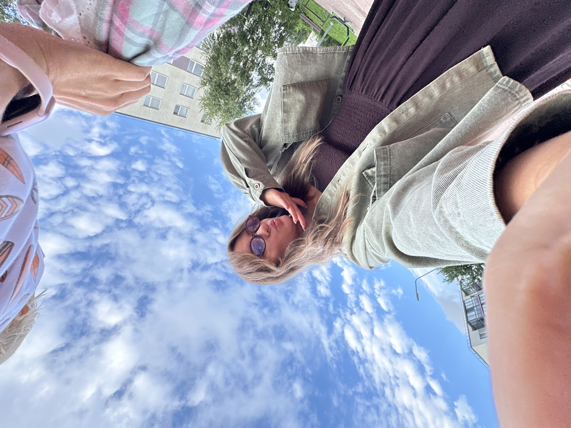 A low-angle, wide-shot selfie shows a young Caucasian woman with blonde hair, wearing glasses, a green jacket, and a dark brown dress, posing with her hand to her face against a bright blue sky filled with scattered white clouds. She is in Vsevolozhsk, Russia, likely outdoors, given the visible sky and background elements of a building, trees, and a street lamp. Part of another person's arm wearing a plaid sleeve and a person's head with blonde hair are visible at the top of the frame. The scene is captured in daylight.