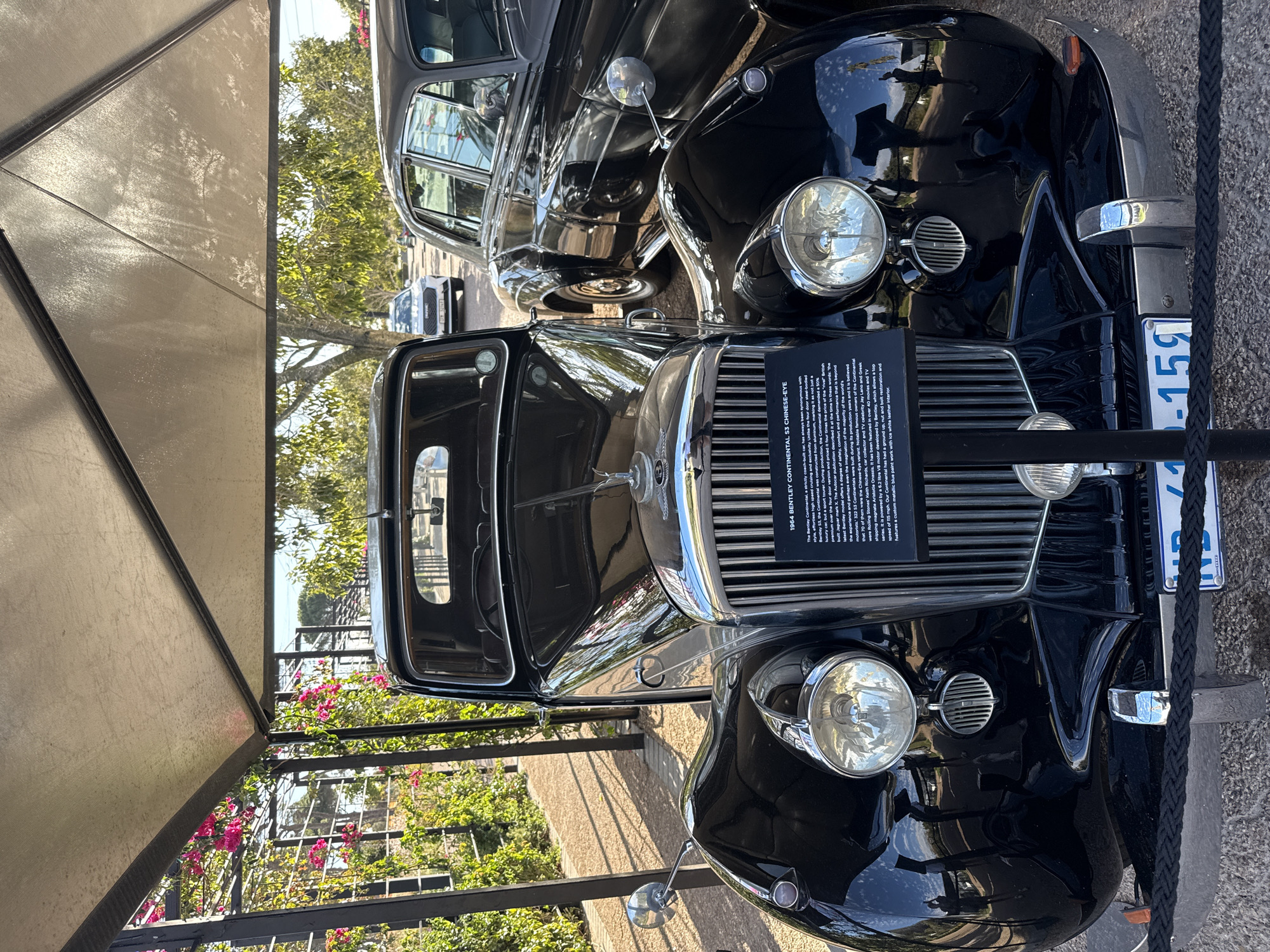 This image shows a 1964 Bentley Continental S3 Chinese-Eye on display, likely at a car show or museum in Stellenbosch Nu, South Africa. The car is black and in excellent condition, featuring a prominent chrome grill and headlights. A sign in front of the car provides details about its history and features. Another classic black car is partially visible behind it. The scene is outdoors, possibly under a canopy or awning, with a glimpse of greenery and pink flowers in the background. A black rope barrier runs along the front of the car. A license plate is visible, reading "NVD 1-159".