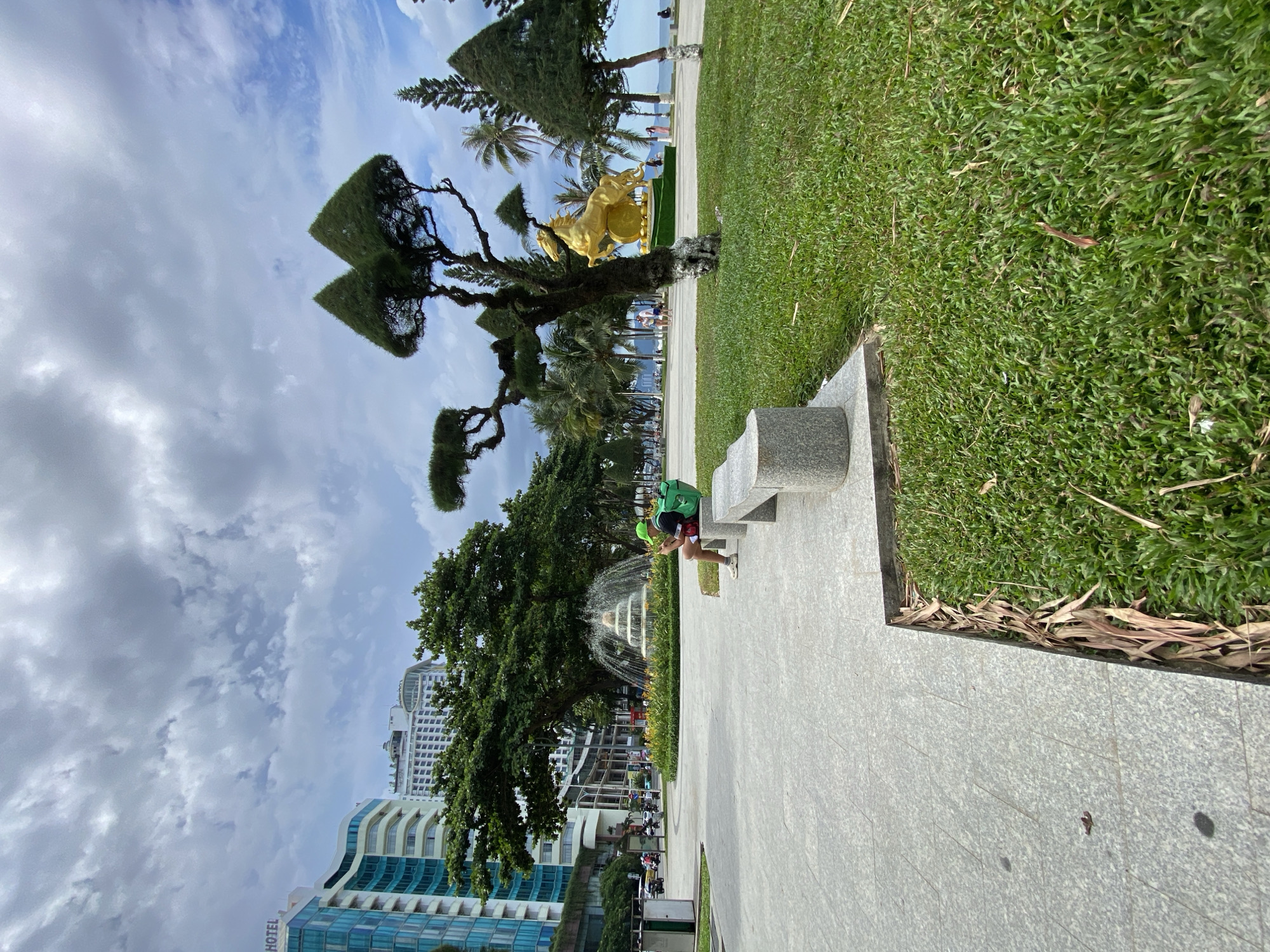The image depicts an outdoor scene with a person sitting on a park bench, holding a phone and wearing a green backpack. The setting appears to be a public park with a paved walkway, lush green grass, and well-maintained trees. In the background, there's a fountain spraying water and a glimpse of city buildings, including what looks like a hotel, suggesting an urban environment. The sky is partly cloudy, indicating a day with mixed weather. The overall scene conveys a sense of relaxation and everyday activity in a park. The text "HOTEL" is visible on one of the buildings. The perspective of the image is slightly tilted.