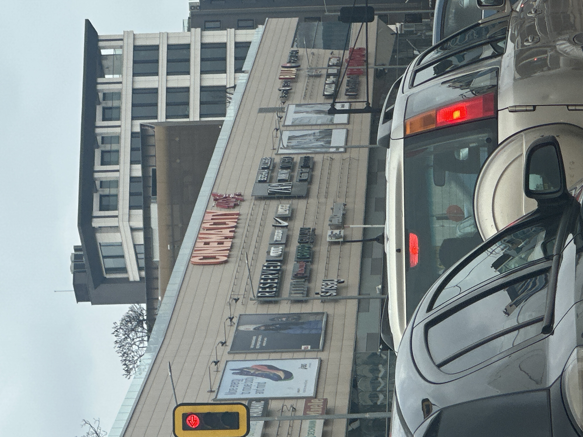 The image shows an urban scene in Sofia, Bulgaria, with a focus on the facade of the Arena Cinema City mall. The facade displays various store signs, including Cinema City, Reserved, Nike, and Sinsay, as well as large advertisements. A multi-story building stands next to the mall. Traffic signals are visible, showing a red light. Several cars, including a silver van and a dark-colored SUV, are in the foreground, suggesting a traffic jam. The sky appears overcast.
