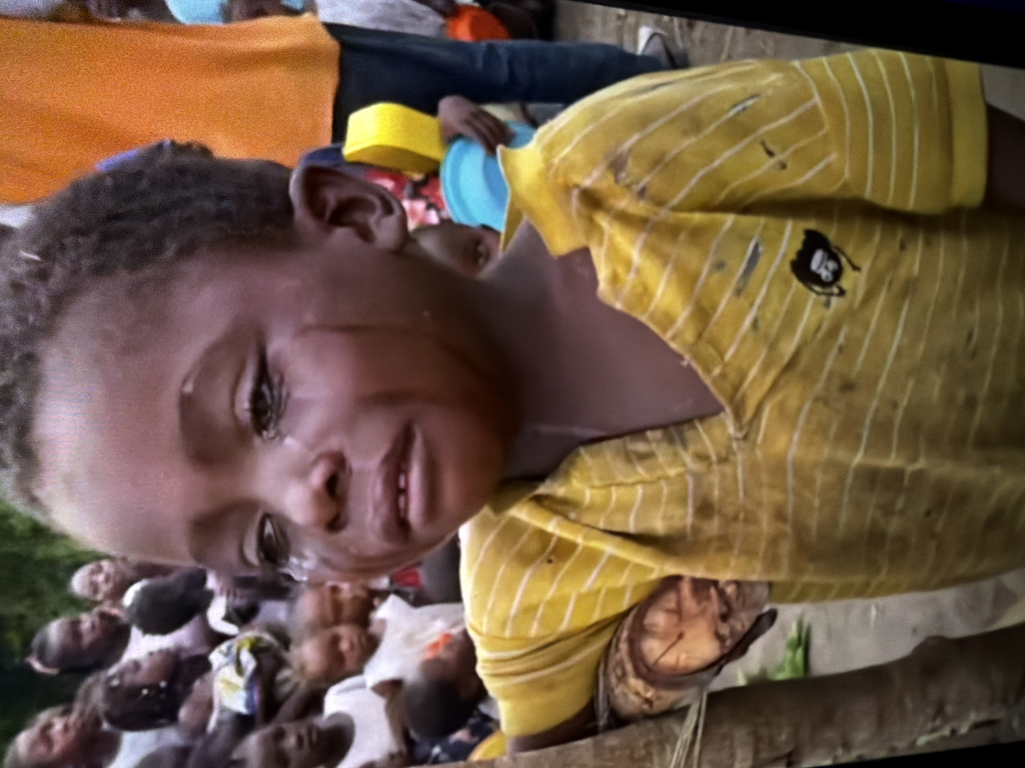 A medium shot shows a young Black child crying, with tears streaming down their face, in an outdoor, possibly rural, environment. The child is wearing a yellow t-shirt with white stripes and a small black graphic on the left side of the chest. The child's left arm is resting on a wooden pole or log. In the blurred background, several other people, including children, are visible. Some are wearing white shirts, and one person in the background is wearing an orange t-shirt. Two plastic containers, one yellow and one blue, are held by individuals in the background. The overall lighting suggests daytime.