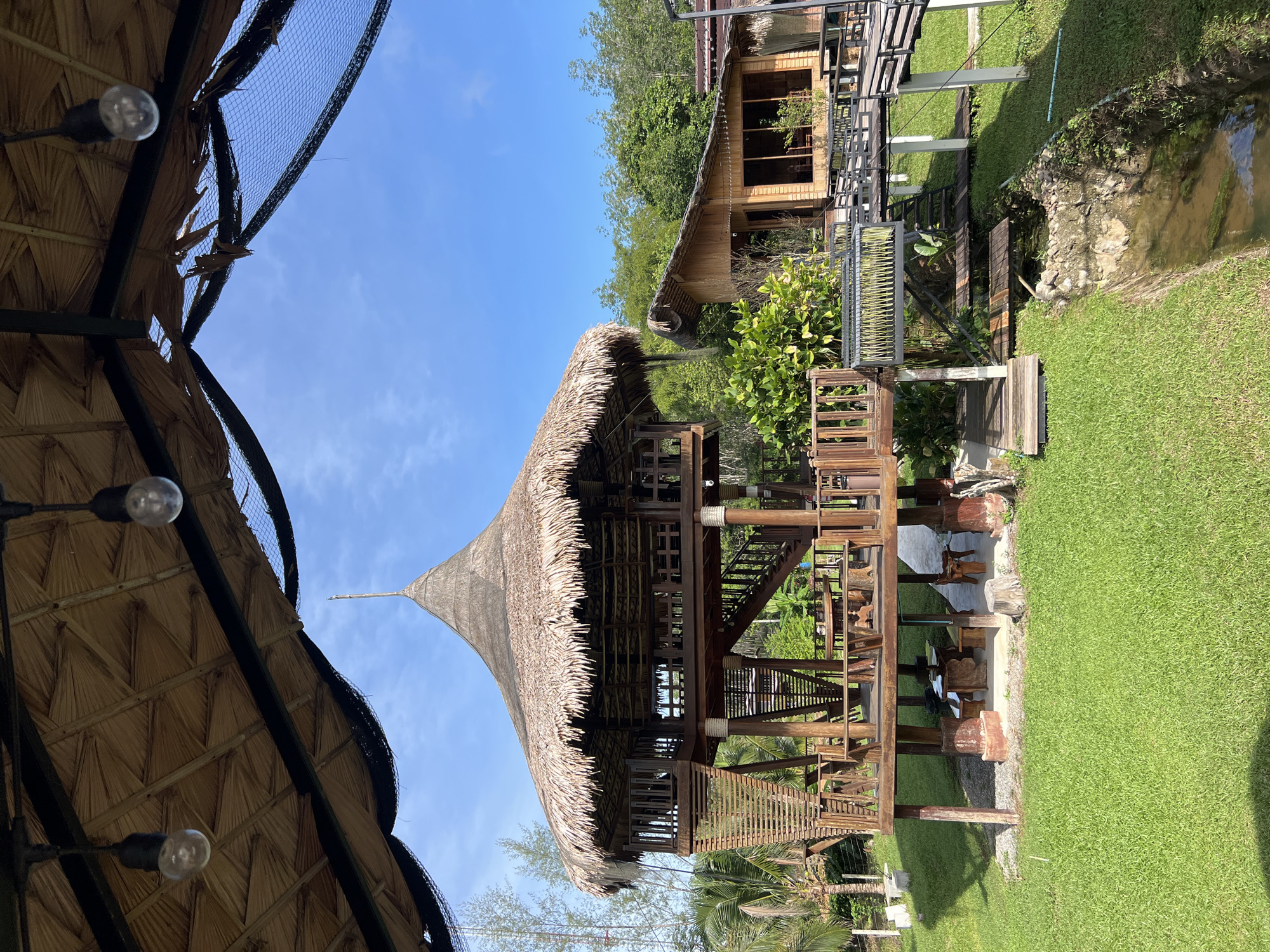 The image depicts an outdoor scene with traditional wooden structures and lush greenery. In the foreground, a thatched-roof pavilion with a conical design is prominent. This structure features intricate wooden latticework and multiple levels, suggesting it might be a seating or dining area. Hanging lights are visible under the thatched roof, indicating it is designed for use both during the day and at night.
Behind this pavilion, another wooden building with a pitched roof and large windows can be seen, surrounded by trees. A wooden walkway or bridge connects different parts of the area, and a small stream or water feature runs through the grassy landscape. The overall setting appears to be a resort, garden, or recreational area, with a focus on natural materials and a tropical ambiance.
The scene is bathed in bright sunlight, suggesting it is daytime. The sky is clear and blue with a few wispy clouds, indicating fair weather. The presence of well-maintained grass and tropical vegetation points towards a warm climate. The location is identified as Tambon Pa Kae Bo Hin, Thailand, which aligns with the architectural style and vegetation visible in the image. No people are present in the frame. The overall mood is serene and inviting, evoking a sense of relaxation and connection with nature. There is no visible text in the image.