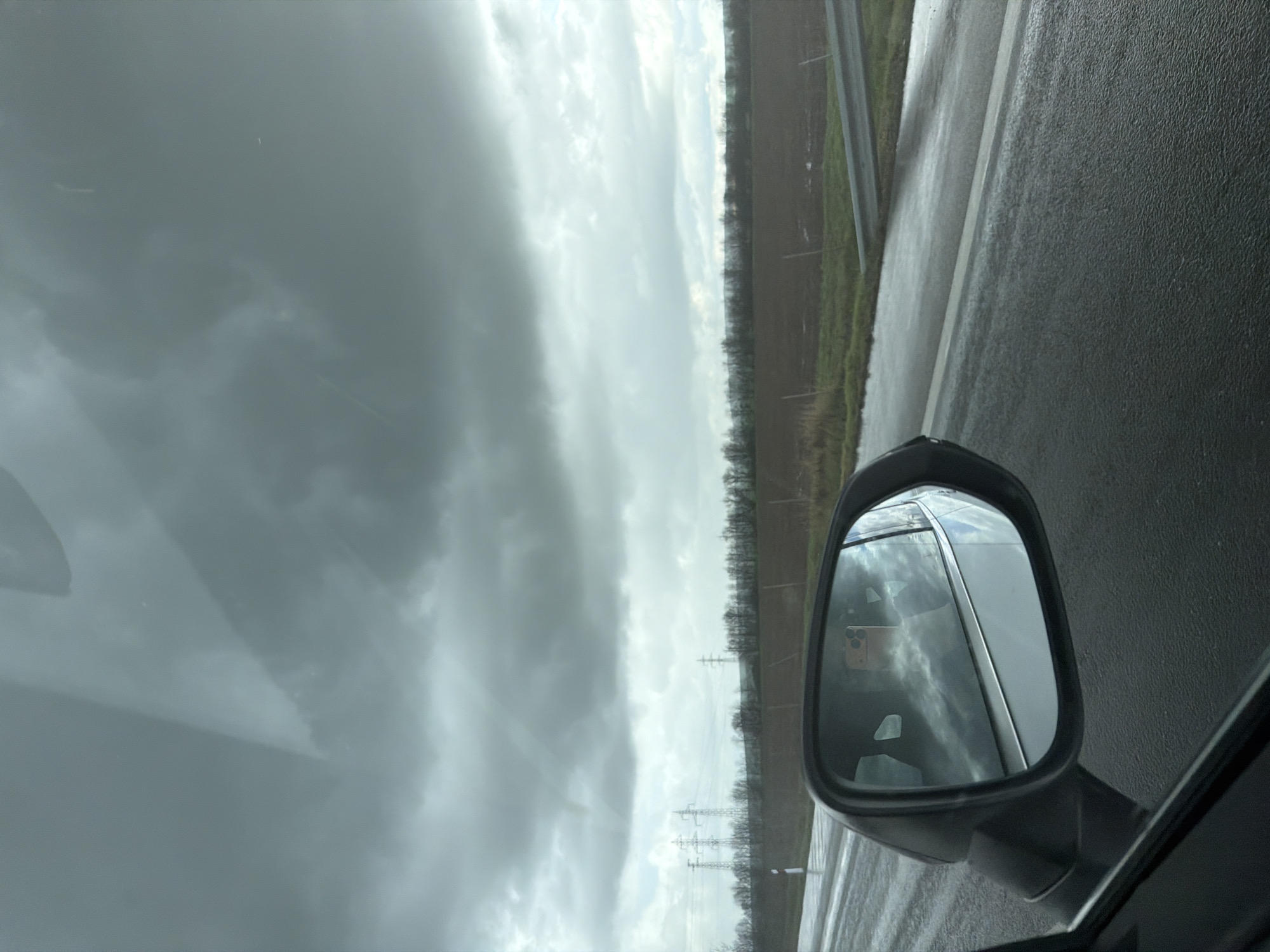A view from inside a vehicle, likely a car, on a road in what appears to be a rural area under a cloudy sky. A side mirror is visible, reflecting the sky and possibly the vehicle. A strip of trees and fields borders the road. There are power lines visible in the distance. There is no indication that the photo was taken in the city of Vodenichane, Bulgaria. No people or subjects are directly visible. The environment consists of the inside of a vehicle, a wet road, fields, and cloudy sky. The implied action is driving. No interactions are visible.