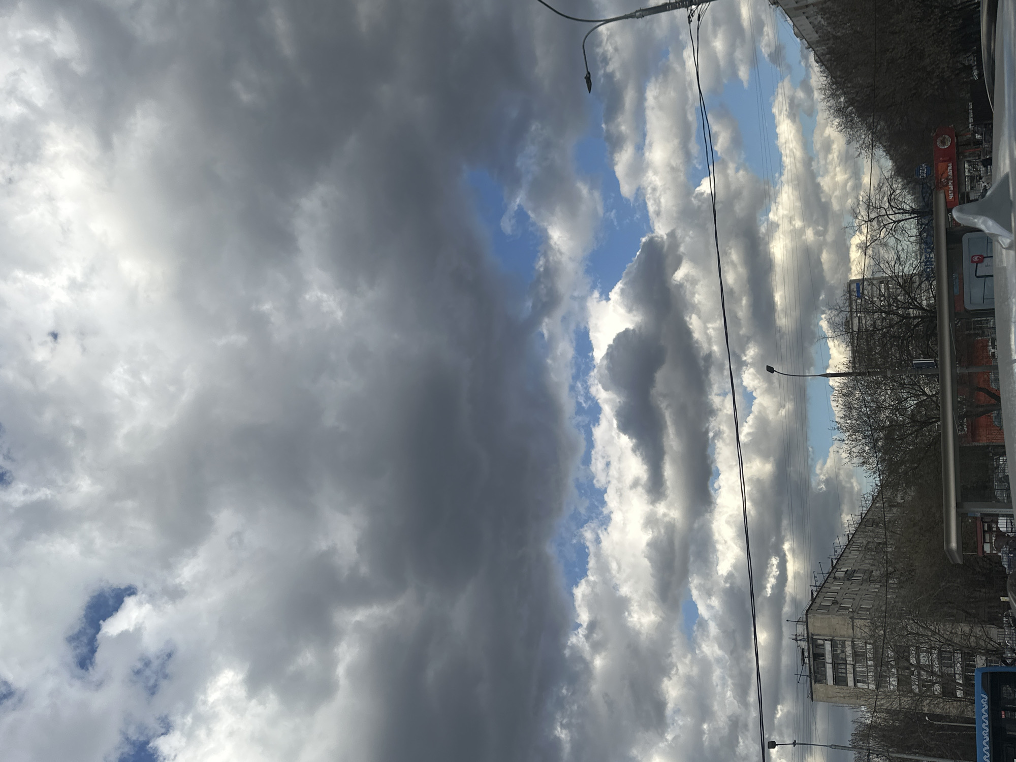 The image presents a view of a Moscow, Russia street scene, focusing on the sky. Overcast conditions prevail, with heavy, dark gray clouds dominating the left side of the frame. A narrow strip of blue sky with lighter, white clouds is visible toward the right. Infrastructure elements such as power lines and street lamps are present. Buildings line the lower right, some obscured by bare trees. A sign written in Russian is also visible.