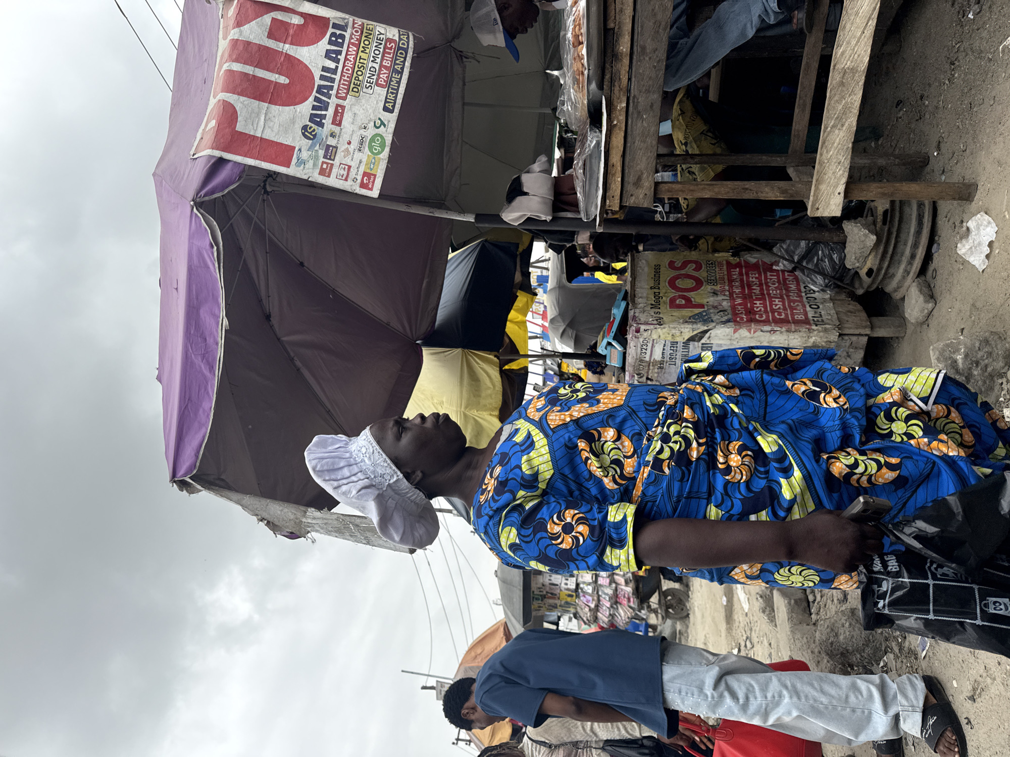 In the city of Lekki, Nigeria, a woman stands facing right, wearing a blue and yellow patterned wrap dress and a white head covering. She holds a black plastic bag in her left hand. Behind her, to the left, a person in a blue shirt and light-colored pants stands facing away from the camera. To the right of the woman is a market stall with a purple canopy. A sign on the stall, partially obscured, has red lettering that reads "FUS" and lists services like "WITHDRAW MONEY," "DEPOSIT MONEY," "SEND MONEY," "PAY BILLS," and "AIRTIME AND DATA." There are other market stalls and people visible in the background, suggesting a busy outdoor market environment. The ground is dirt and debris. The sky is overcast.