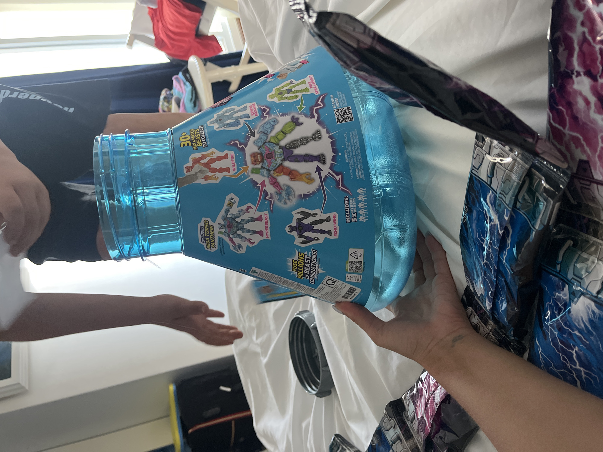 The image shows a close-up of a person's hand holding a large, translucent blue toy container. The container has graphics depicting various toy figures and text. The person's arm is visible, and they are wearing a dark-colored bracelet. In the foreground, there are several sealed toy packages with blue and purple lightning bolt designs. In the background, out of focus, a person's legs are visible wearing dark shorts and a blue t-shirt with white text. There is also a white stool with folded clothes and a white wall behind it. The scene appears to be indoors, possibly on a bed given the white fabric in the foreground. The exact location is unknown.