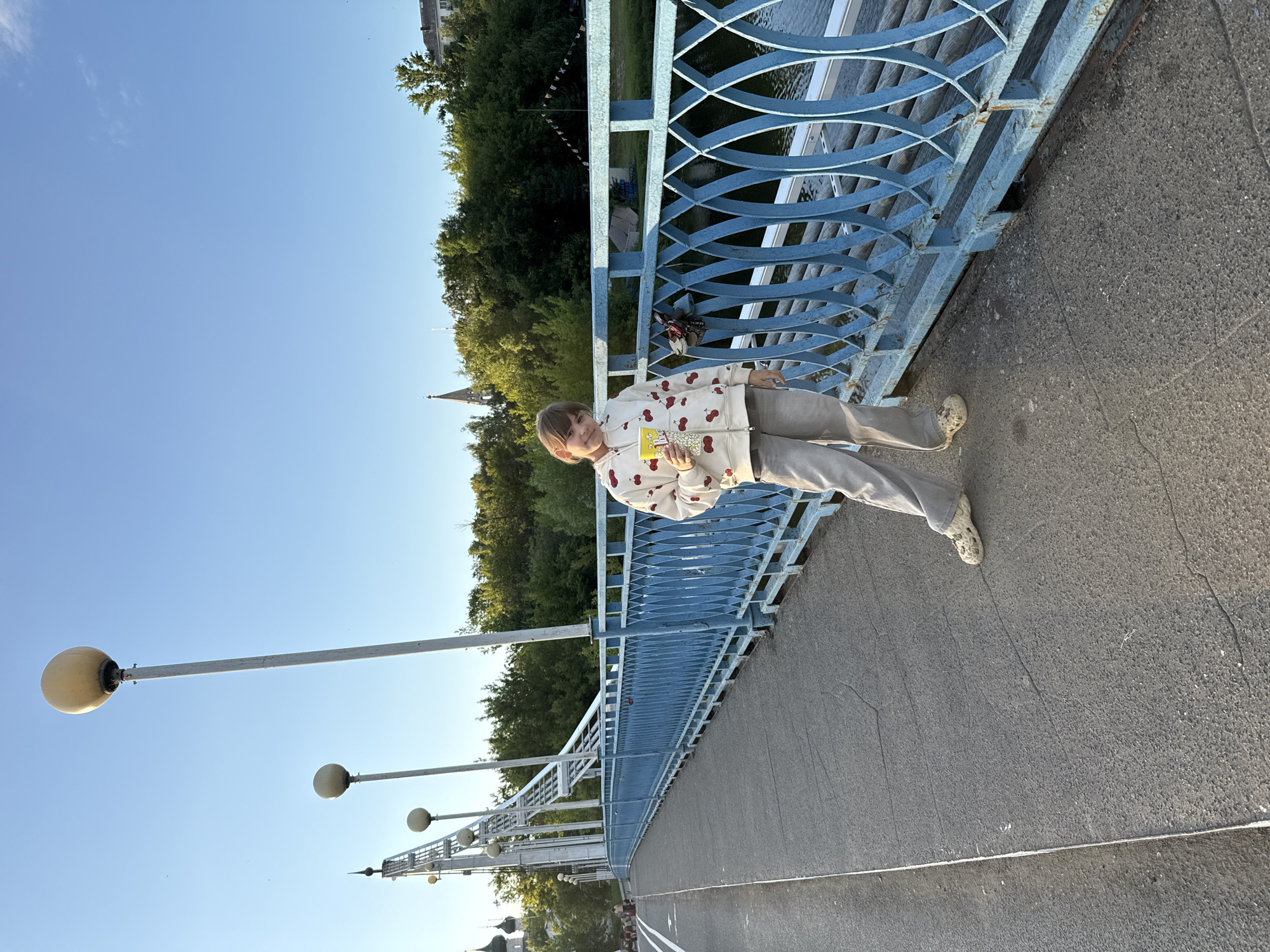 A young person, likely a child or pre-teen, stands on a bridge in Заволжье, Russia, captured in a full-length, daytime portrait. They have light brown hair and are wearing a cream-colored hooded sweatshirt with a red cherry pattern, light grey pants, and patterned clogs. The individual leans casually against a blue metal bridge railing with an intricate, woven-like design. In their hands, they hold a yellow container of popcorn, clearly displaying the text "ВЕСЁЛЫЙ ПОПКОРН" (Cheerful Popcorn).
The scene is set under a clear blue sky with a few faint wisps of clouds, indicative of fine weather. Several white lampposts with spherical light fixtures are visible extending from the bridge structure on the left. Beyond the blue railing, glimpses of dark water are visible, bordered by dense green trees. Distant buildings or spires are partially obscured by the foliage, suggesting a town or city in the background. A small, dark-colored lock is attached to the bridge railing. The bridge's surface is dark grey asphalt or concrete, featuring some cracks and a painted white line. The lighting is bright and even, consistent with late afternoon or early evening.
