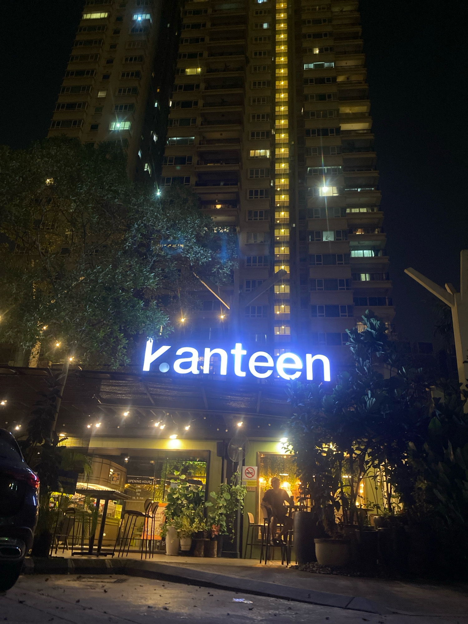 This is a nighttime, ground-level, outdoor shot of a building facade with a neon sign. The building is tall and appears to be an apartment complex or hotel, with many windows lit up. In front of the tall building, at a lower level, is a business with a green facade. A prominent neon sign reads "kanteen" in blue, illuminated letters. There are strings of warm-toned lights draped across the front of the lower business. Trees are visible in front of the business, some with dark green foliage, and some with lights reflecting off their leaves. The ground in the foreground appears to be paved, with some debris or leaves scattered on it. There is a dark-colored car parked on the left side of the frame. Inside the business, through the large windows, there are tables and chairs, and several plants are visible. A person is seated at a table inside the business, silhouetted against the interior lighting. The overall ambiance is that of an urban evening scene, possibly a restaurant or cafe in Kuala Lumpur, Malaysia, given the architectural style.