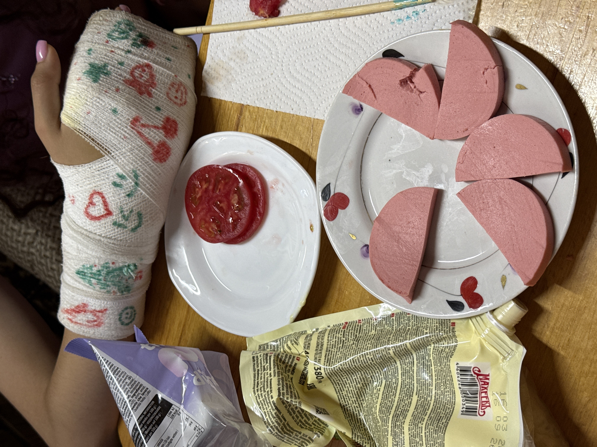 An individual with a bandaged left hand rests on a wooden surface. A pink nail polish is visible on the thumb. The bandage is decorated with printed hearts, Christmas trees, and teddy bears. Beside the hand are two plates of food. One holds two slices of red tomato. The other, a larger plate, contains four half-round slices of cooked sausage. Nearby is a packet of "Makers" mayonnaise written in Cyrillic text. Some candy package is visible. A wooden chopstick and paper towel are in the image. The location is a domestic setting, implied by the informal arrangement of food and the wooden table surface, but there's no specific indicator in the image to confirm it is in Tashkent, Uzbekistan.