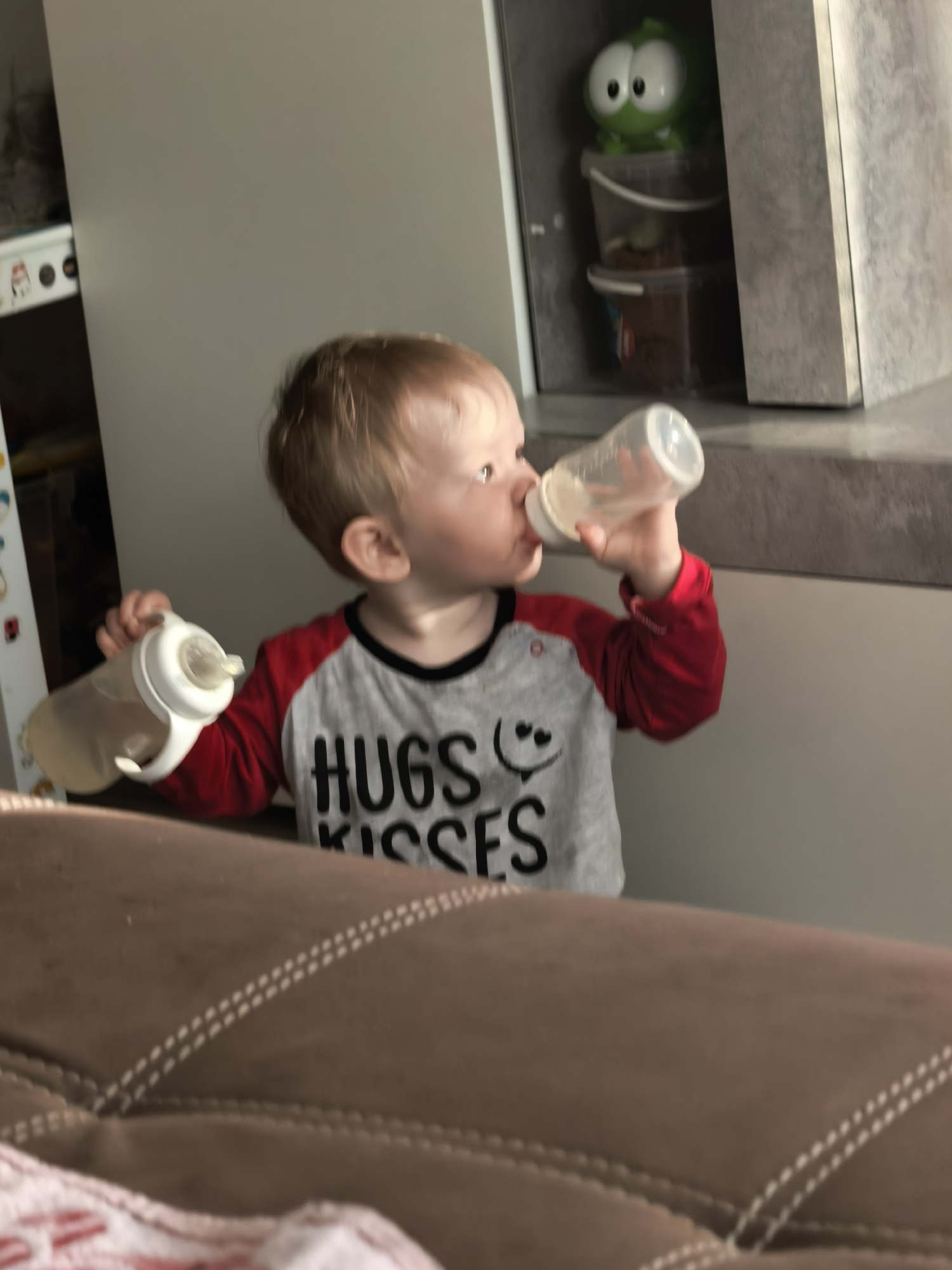 A fair-skinned toddler with light-brown hair is drinking from a baby bottle, while holding another bottle in their other hand. They are wearing a grey shirt with red sleeves and the words "Hugs Kisses" printed in black. The location is an interior with a modern kitchen in the background, visible is a cabinet with a green cartoon figure on the shelves. The toddler is standing in front of a brown couch. The location in the city of Sterlitamak, Russia cannot be determined.