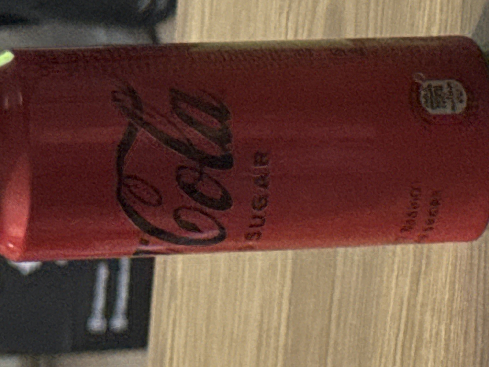 The image is a close-up, slightly blurry shot of a red Coca-Cola can. The can is upright and partially visible, with the Coca-Cola logo clearly discernible in black script. Below the logo, the word "SUGAR" is also visible. There is a small circular emblem near the bottom of the can. The can is placed on a light-colored, textured surface that appears to be a wooden table. In the blurred background, a dark, rectangular object with two horizontal white lines is visible. The lighting suggests an indoor environment. There is no information in the image to indicate the location is Tbilisi, Georgia.