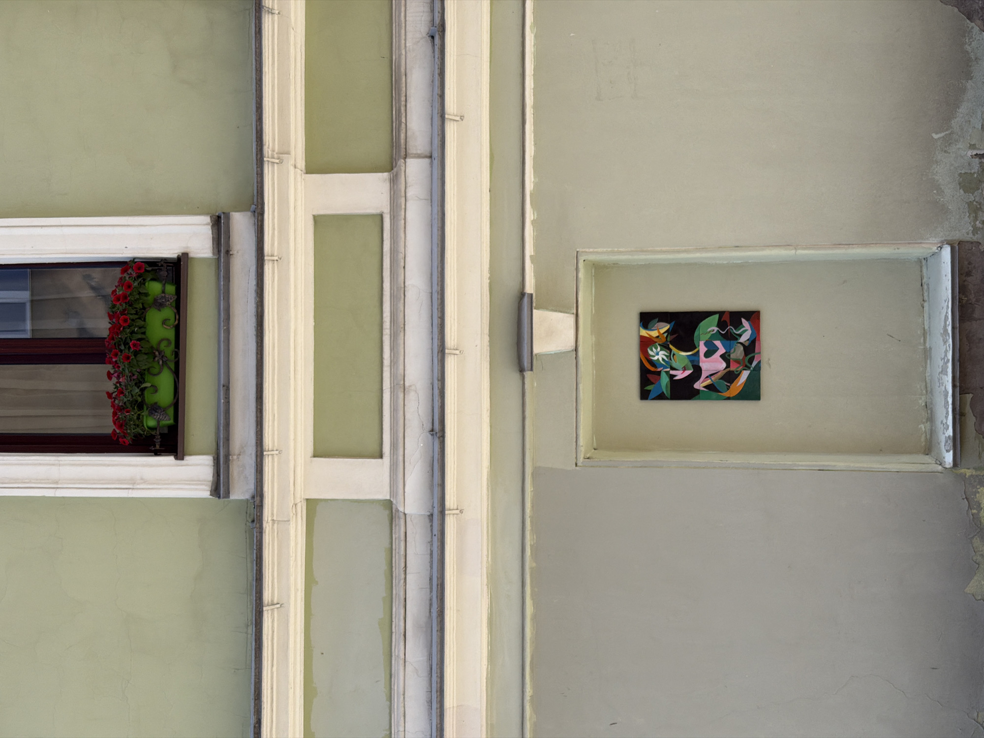 This media file presents an abstract, close-up view of a building facade in Saint Petersburg, Russia, dominated by muted tones of green, cream, and gray. The composition is highly geometric, emphasizing vertical and horizontal lines.
On the left, a portion of a window is visible, featuring a dark frame and white trim. A window box positioned below the window sill contains a lush display of red flowers, possibly petunias, in a green planter. To the right of the window, the facade is adorned with prominent cream-colored architectural moldings that run vertically and horizontally, creating a structured, grid-like pattern. A thin, dark pipe or conduit runs vertically alongside some of these moldings.
On the right side of the image, a rectangular recessed niche, framed by light-colored trim, is set into the wall. Inside this niche, a small, vibrant abstract painting is displayed. The artwork features an energetic arrangement of shapes and lines in various colors, including green, pink, blue, yellow, red, and white, set against a black background.
The building's walls exhibit signs of age and weathering, with subtle cracks and areas of discoloration, particularly noticeable in the upper and lower right corners. The lighting is soft and diffused, suggesting an overcast day or indirect light, casting no harsh shadows. There are no people visible, and no specific activities are depicted. No text is visible in the image.