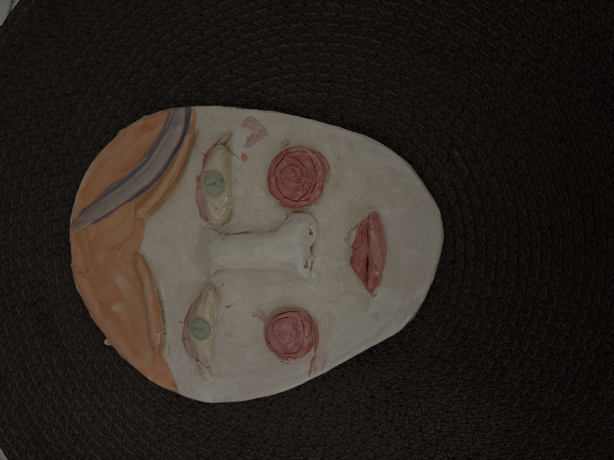 A handmade clay mask with painted facial features rests on a dark, textured surface. The mask has a pale face, protruding nose, and painted eyes with green pupils. There are two pink rose-like shapes representing cheeks and red markings for the mouth and forehead. The left side of the mask has orange hair with a blue stripe. The dark, textured surface appears to be a woven material. There is no indication of a location in Saint Petersburg, Russia.