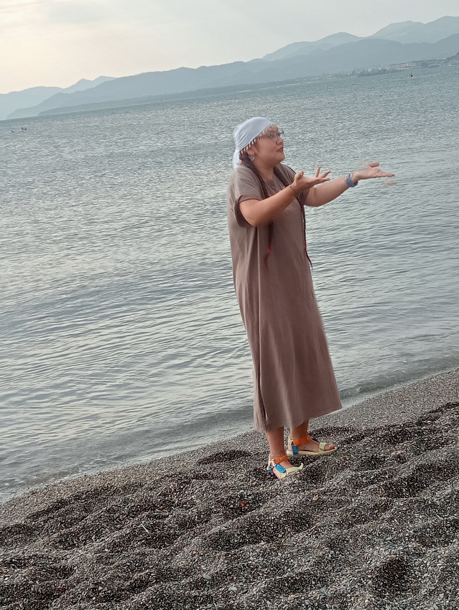 The image depicts a woman standing on a pebble beach, with the sea in the background. The woman is wearing a long, loose-fitting brown dress, a light-colored headscarf, and colorful sandals. She is looking upwards and to the side with her hands outstretched, as if speaking or gesturing. The sea is calm, with gentle ripples. In the distance, a range of hazy mountains forms the horizon. The overall scene is set against a soft, overcast sky. The location appears to be a coastal area, possibly a beach near Fethiye, Turkey, given the mountainous backdrop and the characteristics of the shoreline.