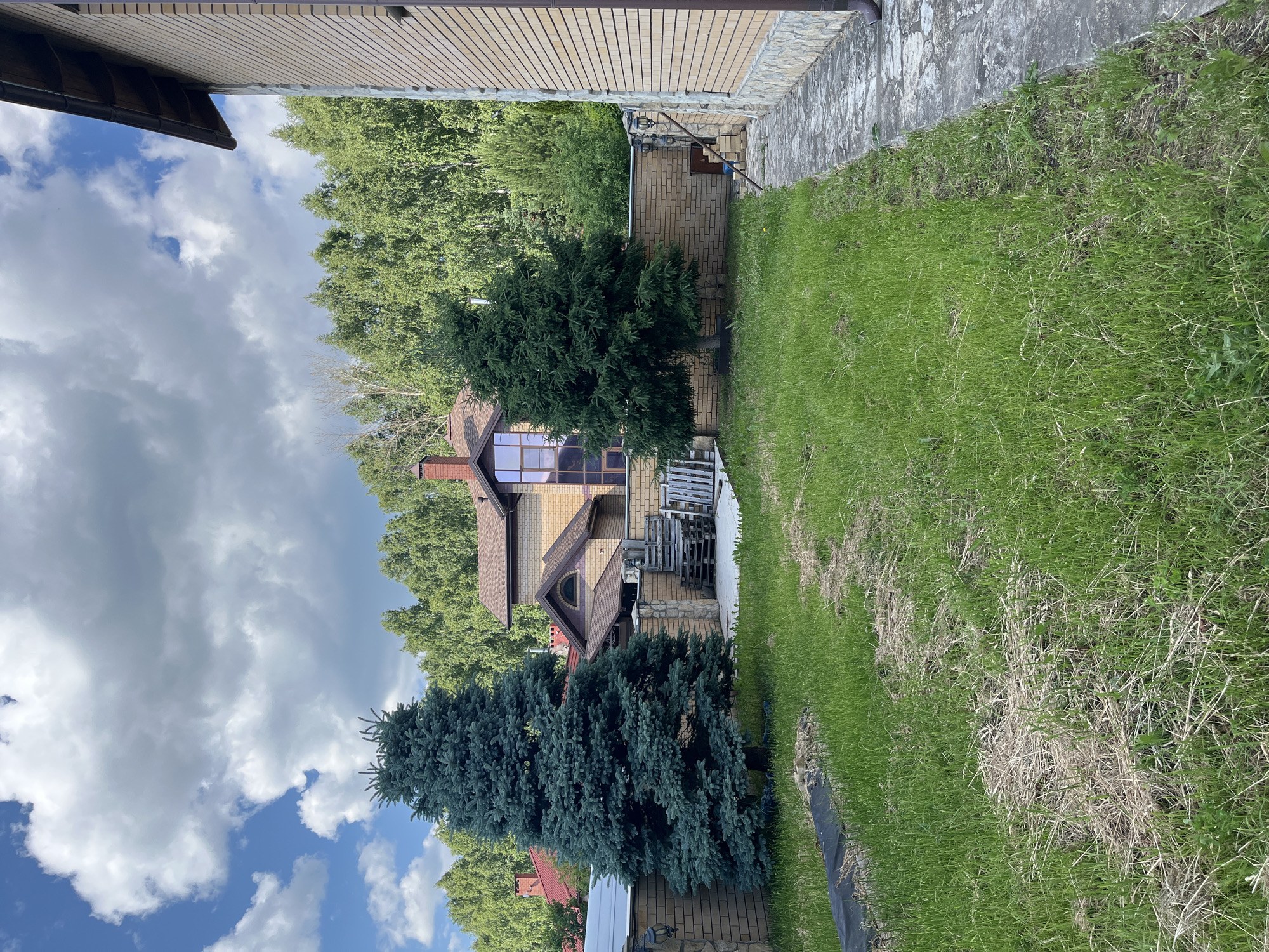 The image depicts a backyard scene with a brick house and lush green lawn in the foreground. Two prominent coniferous trees, one a blue spruce, stand tall in the yard. A low brick wall surrounds a patio area with wooden pallets and stairs. In the background, a dense forest of deciduous trees forms a natural backdrop. The sky is partly cloudy with blue patches visible. The location is identified as Orekhovka, Russia.