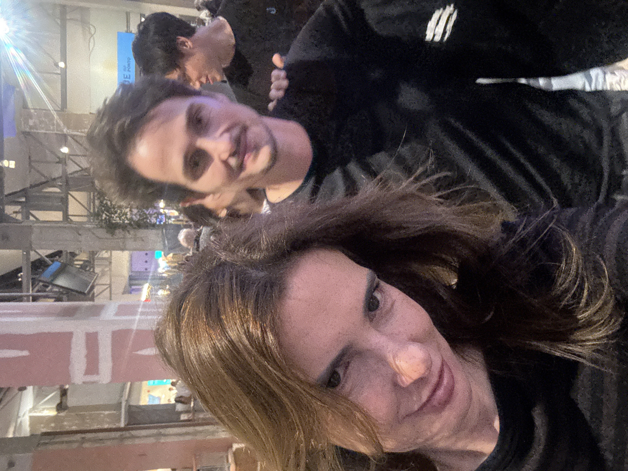 The image shows a selfie of a woman with shoulder-length brown hair, wearing a black top. Behind her and slightly above is a man with short brown hair, also in a black top featuring a white logo on the shoulder. In the blurred background, there are architectural elements suggesting an indoor space with exposed infrastructure, possibly under construction or renovation, including scaffolding and lighting fixtures.
Given the location of Vila Nova de Gaia, Portugal, this could be an event or gathering taking place indoors, possibly in a renovated warehouse or event space. The bright light in the background could be stage lighting.