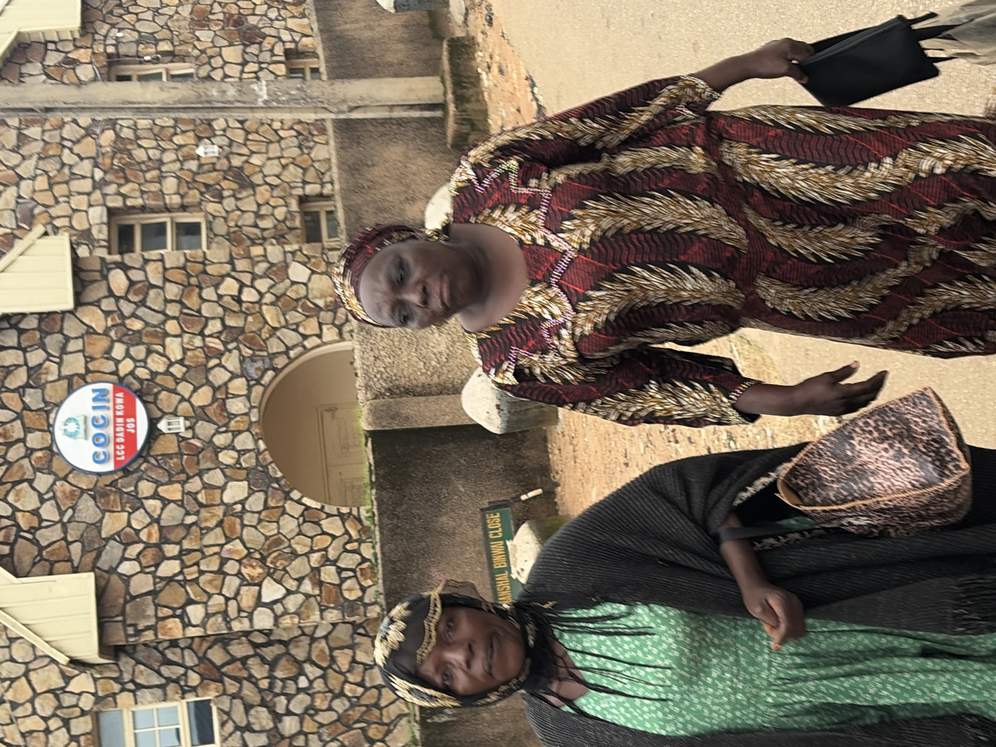 The image shows two women standing outside a stone building with a sign that reads "COCIN LCC BADIN KOWA JOS." The woman on the left wears a green dress and a black headscarf, while the woman on the right wears a patterned red and gold dress and a matching headscarf. The building appears to be a church or administrative office, with arched doorways and windows. A street sign in the foreground indicates "MANSHAL BINWAI CLOSE," suggesting the location is in Jos, Nigeria. The ground is a dirt or gravel path.