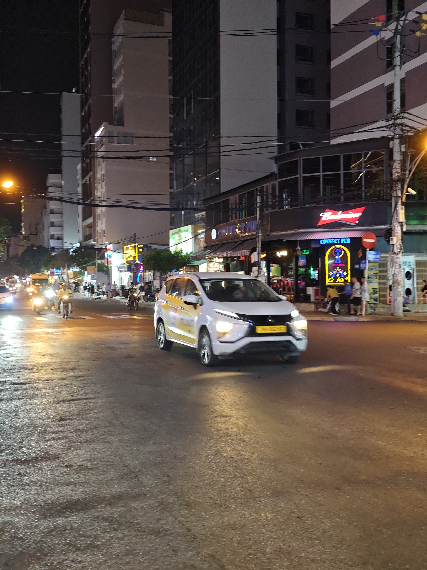 The image depicts a street scene at night in Nha Trang Ward, Vietnam. A white car with yellow accents and a license plate is in the foreground, appearing to be in motion. The street is wet and reflects the lights from the buildings and streetlights, creating a glossy effect. On the right side of the road, there is a building with a Budweiser sign and a neon sign that says "CONNECT PUB". Several people are visible on the sidewalk in front of the pub. Further down the street, traffic, including motorcycles and a yellow van, is visible, illuminated by headlights and streetlights. The background consists of tall buildings, suggesting an urban environment. The overall atmosphere is that of a busy, illuminated city street at night.