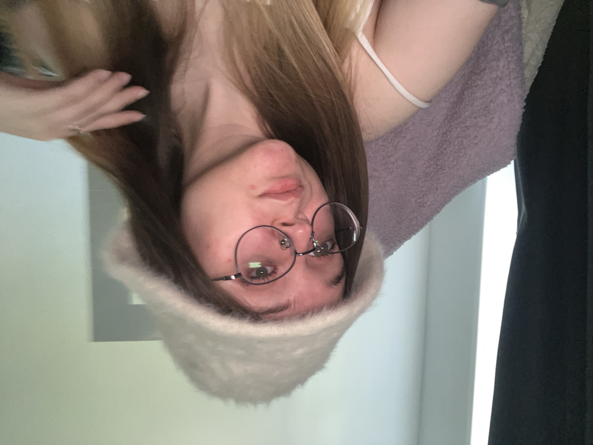 A young woman with light brown hair and glasses wears a white, fuzzy hat. She's also wearing a white tank top and is wrapped in a purple fuzzy blanket. Her right hand is visible near her hair. The background includes a white wall and a dark curtain. The location is not discernible from the image provided, as there is nothing to indicate the location is Pushchino, Russia.