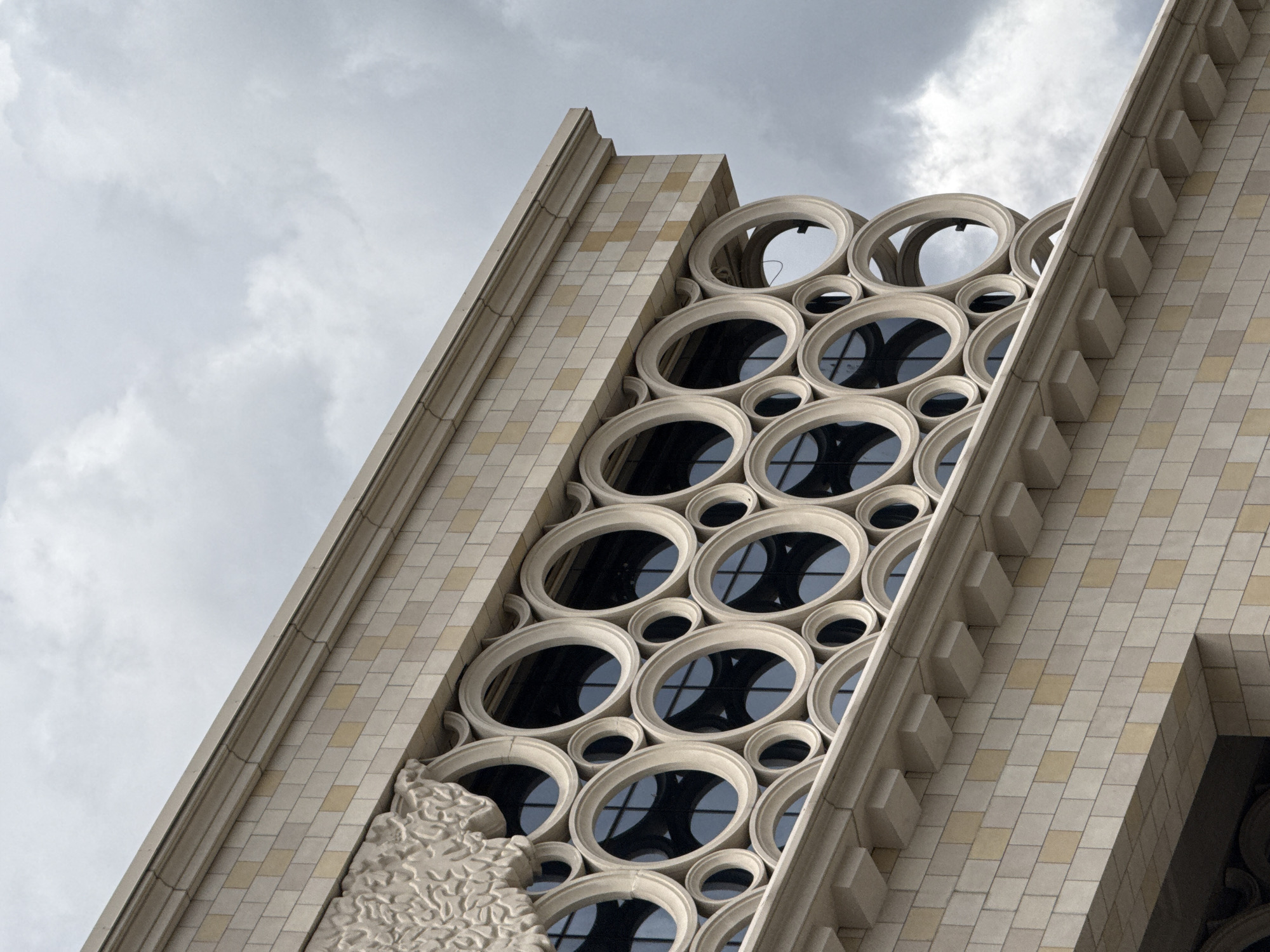 The image shows a close-up of the facade of a building with a unique architectural design. The building is constructed with light-colored stone blocks, some of which are a pale yellow hue. The upper section features a cornice with decorative moldings and a row of projecting rectangular blocks. Below this is a prominent band of circular or oval decorative elements, arranged in multiple rows and varying in size. These circular elements are made of a lighter material and appear to frame windows or openings, which are dark and reflect the cloudy sky. In the upper left corner, a sculpted decorative element with a floral or leafy pattern is visible. The sky is overcast with dark, heavy clouds. The building's style suggests it is located in Moscow, Russia, though the specific location is not identified. There are no people or actions depicted in the image.