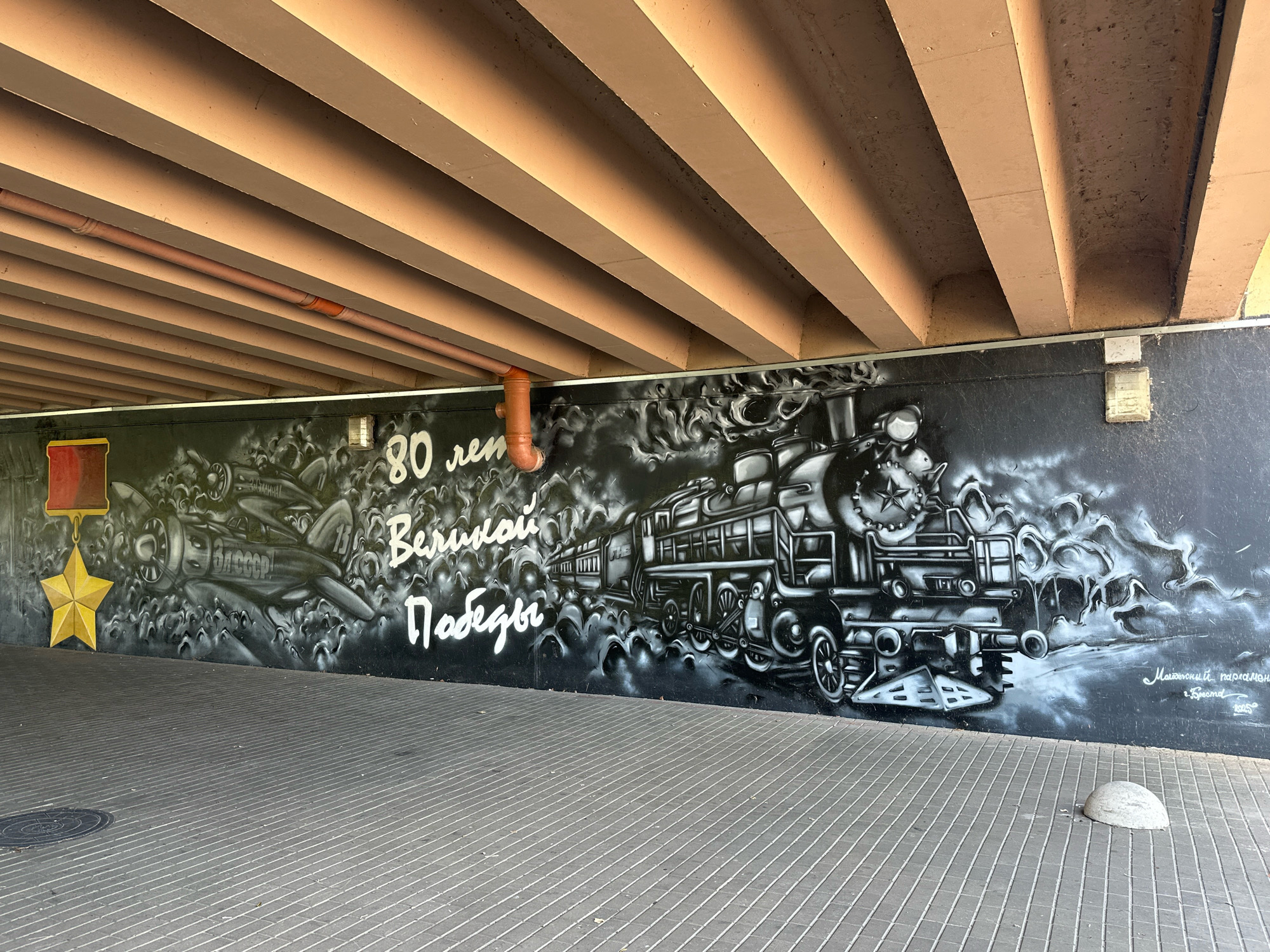 The image shows a mural painted on a wall beneath an elevated structure, likely a bridge or overpass, in Brest, Belarus. The mural features a black and white depiction of a vintage steam train on the right side and an airplane on the left. The train is a prominent subject, rendered with intricate detail, including its wheels, pistons, and steam. The airplane is also detailed, with visible propellers and markings.
Above the airplane, there is a yellow star hanging from a red ribbon, symbolizing a medal. Text in white graffiti-style font is present on the wall. The visible text reads "80 лет Великой Победы," which translates to "80 years of Great Victory." This inscription, along with the imagery of a train and airplane, strongly suggests the mural commemorates Victory Day, a significant holiday in Belarus and Russia.
The environment is urban, with a paved walkway in the foreground. The walkway is constructed of grey rectangular tiles laid in a diagonal pattern. A concrete bollard is visible on the walkway to the right. The underside of the elevated structure consists of parallel beams, painted in a beige or light brown color, with exposed piping running along one of the beams. The overall scene depicts a public space adorned with historical art.