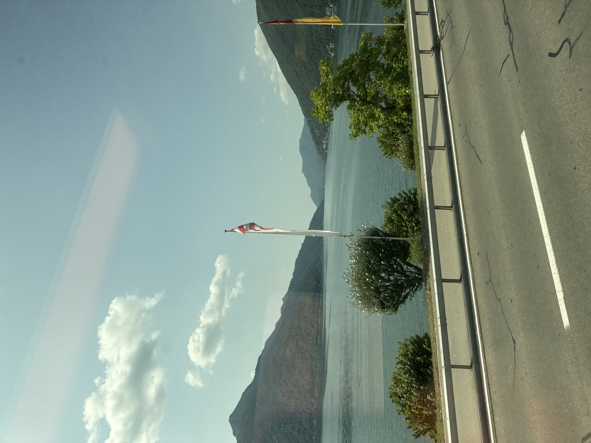 The image depicts a scenic view of Lake Lugano in Bissone, Switzerland. In the foreground, a paved road with white lane markings runs parallel to a metal guardrail. Beyond the guardrail, a body of water stretches out, reflecting the blue sky and scattered white clouds. Along the lakeshore, lush green trees and bushes are visible. Two flagpoles stand near the water's edge; one displays a Swiss flag, and the other a red and yellow flag. In the background, rolling hills covered in dense green foliage rise steeply from the lake. The overall scene is one of natural beauty and tranquility, characteristic of a lakeside town.