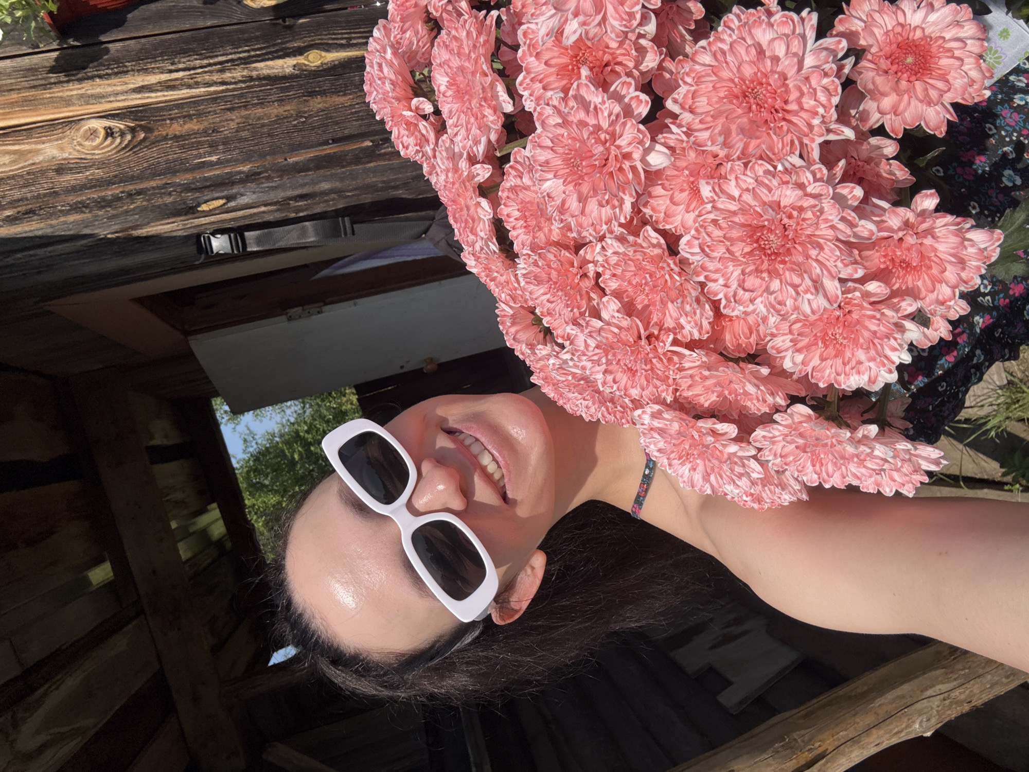 A woman in white rectangular sunglasses and a floral top holds a large bouquet of pink chrysanthemums in front of a rustic wooden building. The building appears to be a shed or outbuilding with weathered wooden planks and a wooden roof structure visible. Sunlight streams down, casting shadows and highlighting the texture of the wood and the petals of the flowers. The woman is smiling and looking towards the camera. The scene suggests an outdoor setting, possibly a rural or semi-rural environment. Zasechnyi, Russia is a village in the Republic of Mordovia, which is known for its agricultural landscape and traditional wooden architecture. Given the context of the image, it is plausible that this scene is located in Zasechnyi, Russia, within the grounds of a dacha or a rural home.