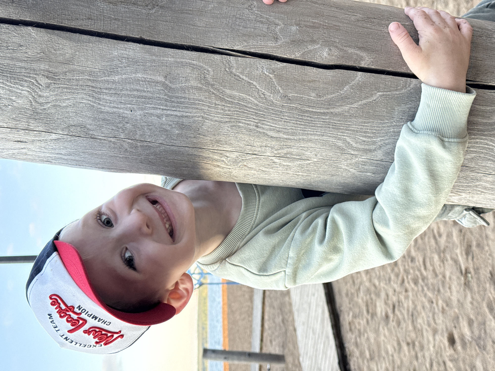 The image, presented sideways, captures a young child smiling brightly while peeking over a large, weathered wooden structure. The child, likely a boy, is positioned such that his head and upper body are seen from below the structure, with his hands gripping the top edge of the wooden planks. He wears a light green long-sleeved sweatshirt and a baseball cap with a red brim, white front, and blue sides. The cap clearly displays the text "New League" in red cursive, flanked by "EXCELLENT TEAM" and "CHAMPION" in smaller white text. His wide eyes and broad smile convey a joyful and playful emotion.
The foreground features thick, greyish-brown wooden planks with visible grain and cracks. In the background, a bright, clear blue sky indicates daytime and fair weather. Below the wooden structure, the ground is sandy, suggesting a beach or outdoor play area. A blurred distant horizon shows faint land or water with indistinct colorful elements. The location context for this scene is Chaikovsky, Russia.