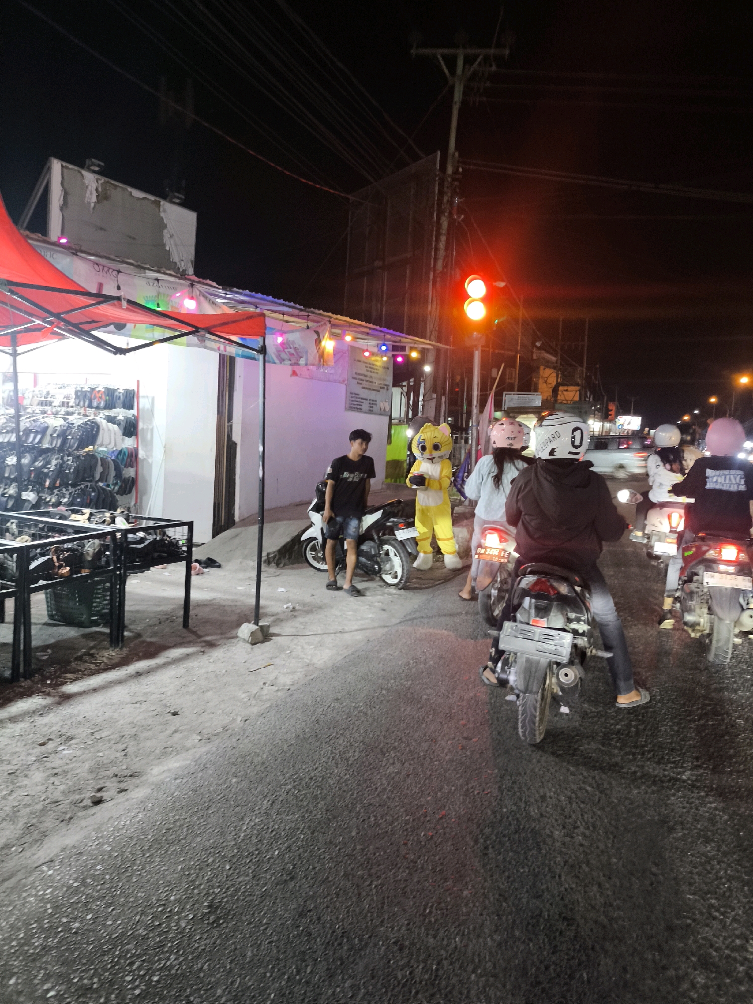 This is a night time, eye-level, medium shot of a street scene in Luwuk, Indonesia. In the foreground, several people on motorcycles are stopped at a traffic light. One motorcycle rider in the center of the frame is wearing a black hoodie and a white helmet. To their left, a person in a yellow cat costume stands on the sidewalk. Behind them, two other people on motorcycles, one wearing a pink helmet, are also stopped. The background shows a brightly lit stall selling shoes and a utility pole with a traffic light. The road is paved and appears wet, reflecting the streetlights and the traffic light.