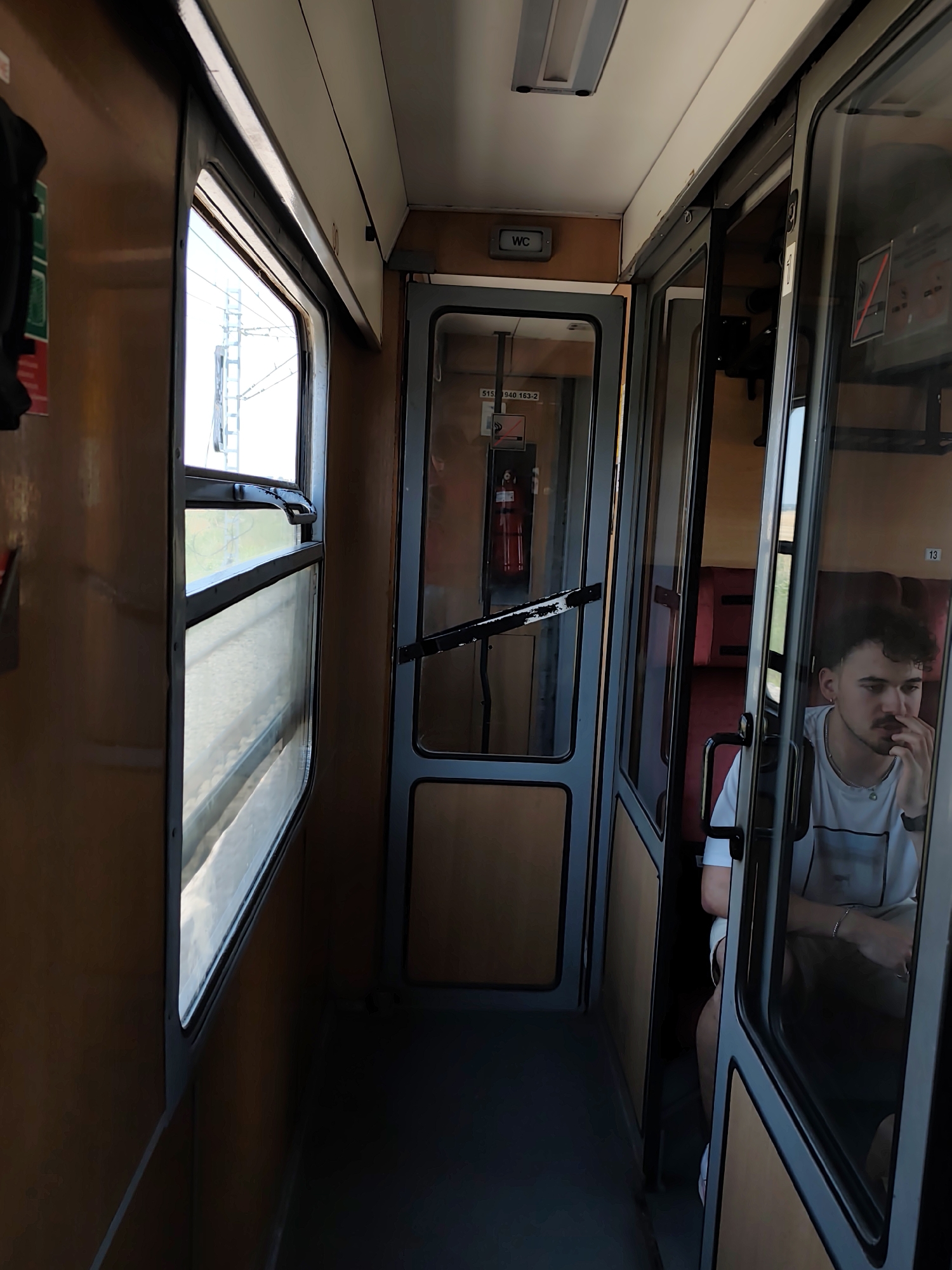 The image shows the interior of a train compartment. A young man with dark curly hair and a mustache is seated in the compartment to the right, looking downwards and resting his chin on his hand. He is wearing a white t-shirt and beige shorts. The train compartment has wooden paneling on the walls and gray doors with glass panels. Through the glass doors, red seats are visible. On the left side of the image, a window shows an outdoor scene with what appears to be electrical pylons, suggesting the train is in motion or passing through a rural or semi-urban area. A sign that reads "WC" is visible on the wall. The lighting suggests it is daytime, with sunlight filtering in through the windows. The overall environment is consistent with a train carriage.