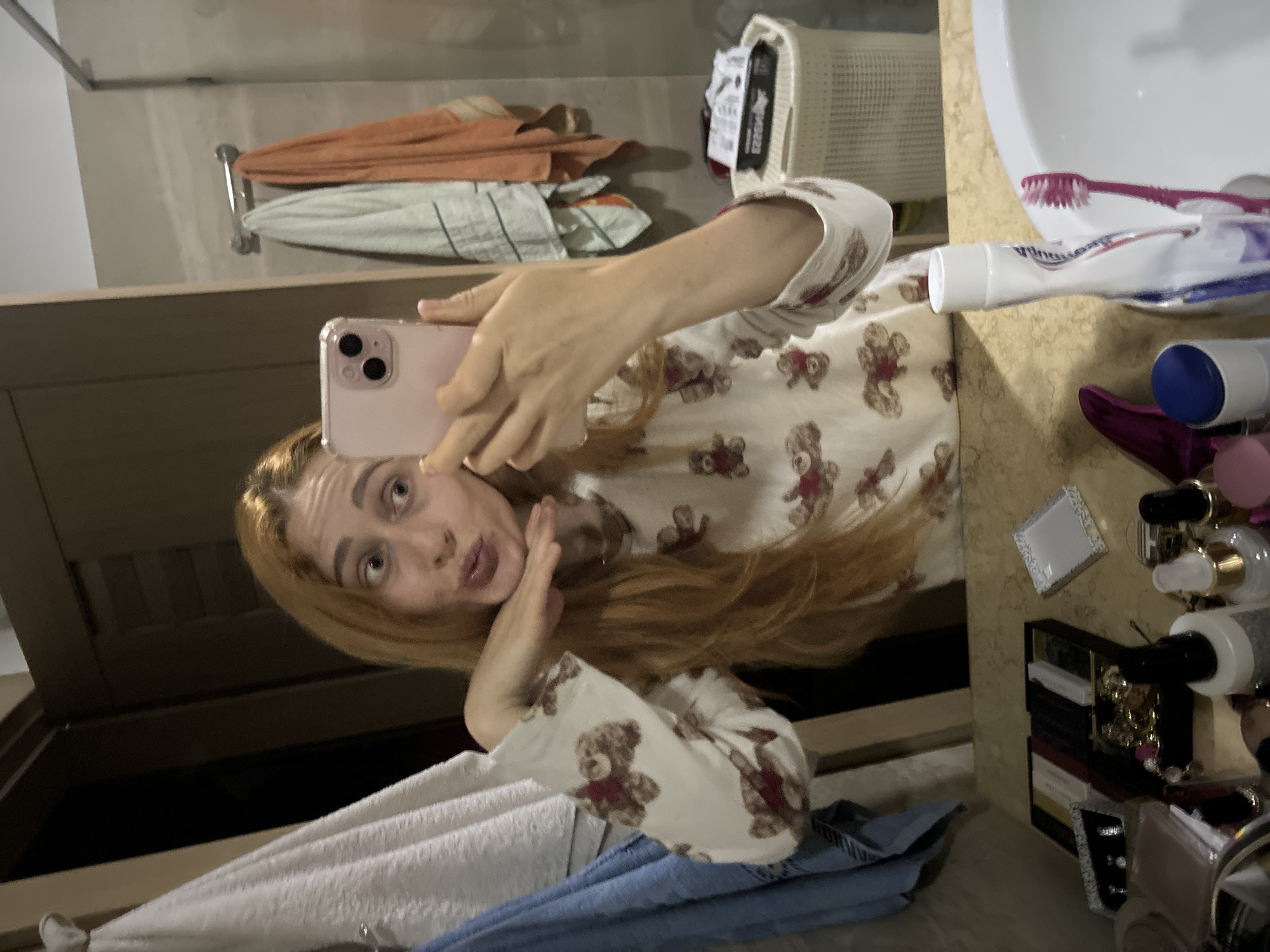 A woman is taking a selfie in a bathroom. The bathroom has towels on a rack, a white hamper, and a sink with toiletries. The woman is wearing a white top with teddy bear print. The location is unknown and cannot be determined.