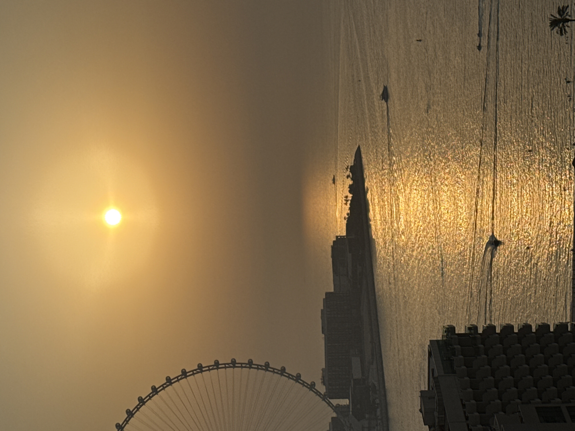 This image captures a sunset scene over the water in Dubai, United Arab Emirates, likely from a high vantage point. A large Ferris wheel, resembling the Ain Dubai, is partially visible on the left side of the frame. In the middle ground, several buildings are silhouetted against the bright sky, suggesting a coastal urban development. The sun is a brilliant orb low in the sky, casting a golden glow across the calm water, with reflections shimmering on the surface. Several boats, including yachts and smaller vessels, are cruising on the water, leaving visible wakes. In the foreground, another building with a distinctive architectural style, characterized by its balconies and patterned facade, is visible. The overall impression is one of a serene and picturesque evening in a modern city.