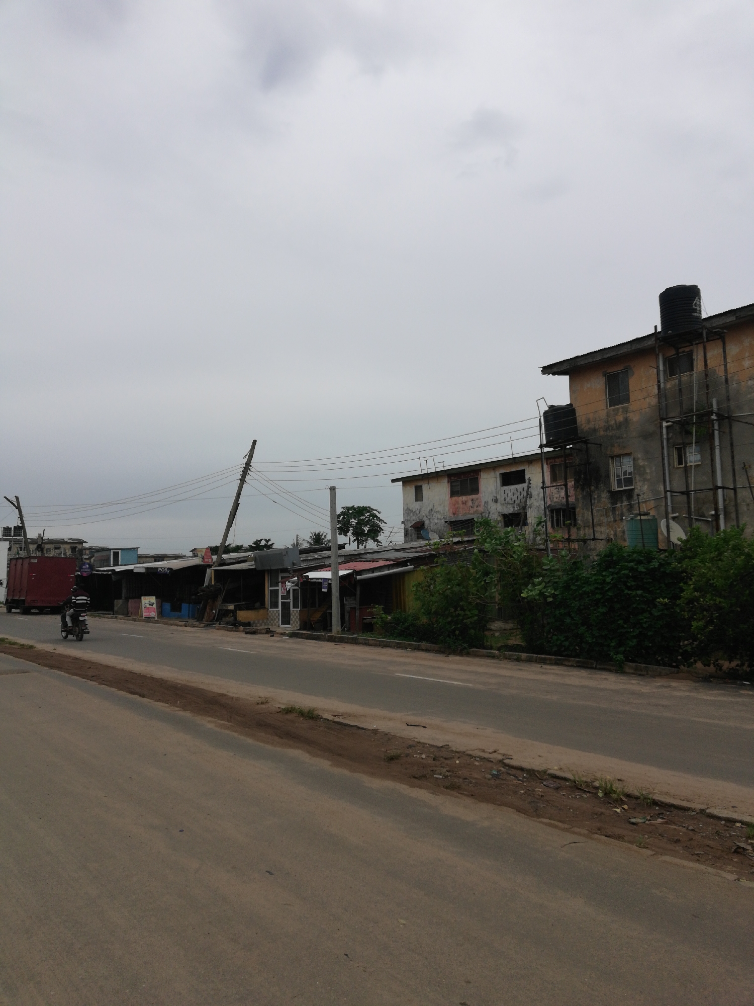 The image depicts an urban scene in Unknown, Unknown, likely a developing area. The sky is overcast. A paved road runs through the frame, showing signs of wear. Along the right side of the road, there's a narrow dirt and debris-filled strip separating the road from the buildings.
A two-story building with visible signs of aging stands on the right. It has water tanks on the roof and external piping running up the side. Adjacent to this building are smaller, informal structures, possibly makeshift shops or residences, with corrugated metal roofs.
On the left side of the road, there are more similar structures and a red truck. Power lines stretch across the frame, connected to leaning utility poles. A motorcyclist is traveling along the road, heading away from the viewer.
