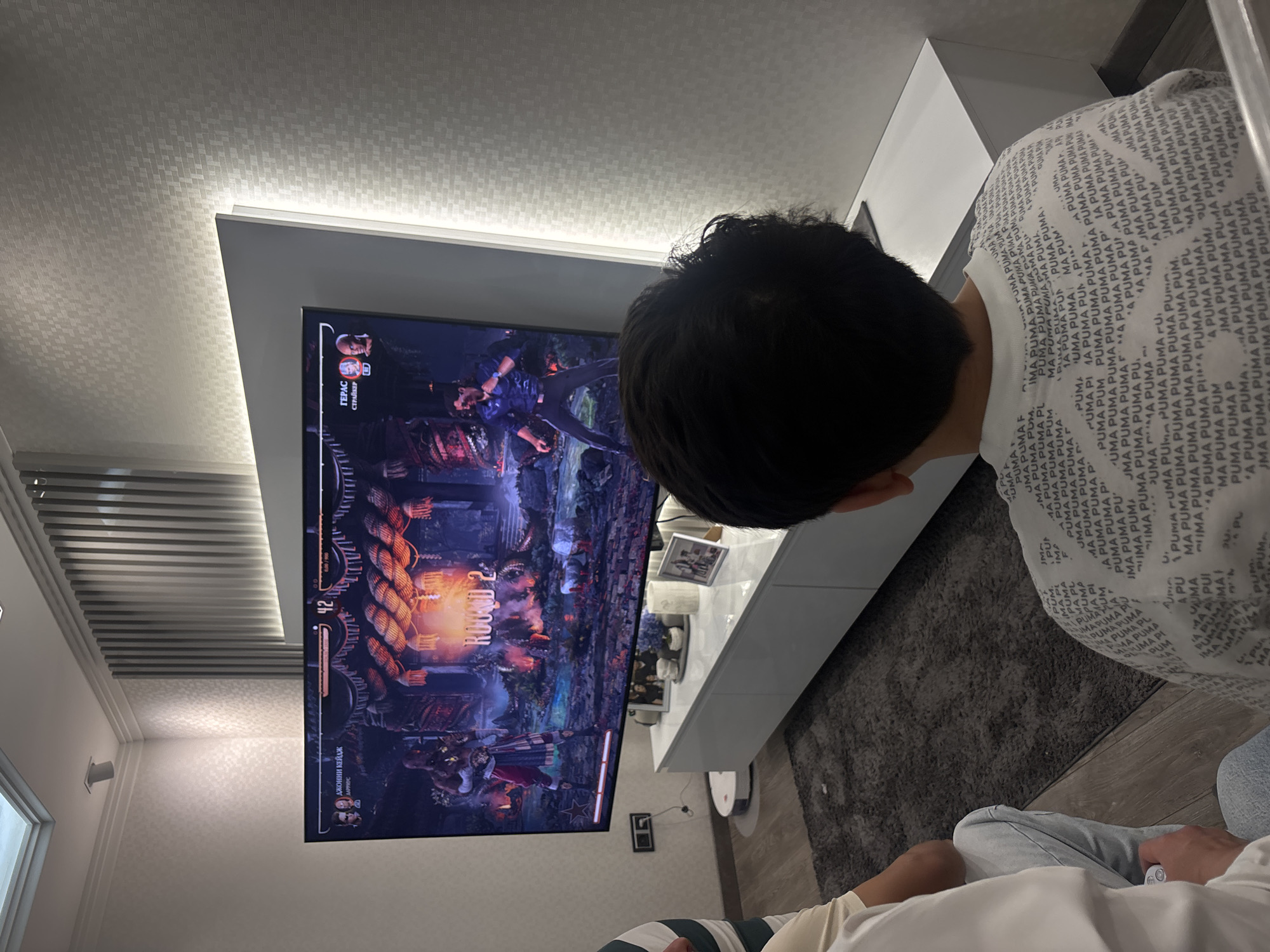 An overhead, slightly angled shot captures a person from behind, deeply engrossed in a video game displayed on a large, wall-mounted television screen in what appears to be a modern living room in Yerevan, Armenia. The person is seated, wearing a white t-shirt adorned with a repeating "PUMA" logo pattern, with their back facing the camera.
The TV screen shows an active fighting game, specifically "Mortal Kombat" given the character names. The game is in "ROUND 2," featuring two characters with health bars visible at the top: "ГЕРАС" (Geras) on the left and "ДЖОННИ КЕЙДЖ" (Johnny Cage) on the right. Below Geras's name, the word "СТРАЙКЕР" (Striker) is visible, likely indicating a special ability or assist character. A score of "42" is visible near the health bars. The in-game environment is dark and detailed, possibly a dungeon or industrial setting.
Below the television, a sleek, white entertainment console or cabinet holds a few decorative items and framed pictures. A robot vacuum cleaner is visible on the light wooden floor beneath the cabinet. The room features a patterned ceiling with indirect LED lighting running along the edge of the wall where the TV is mounted, casting a soft glow. Another section of the ceiling, to the left, has a slatted design with similar hidden lighting. A dark grey rug covers a portion of the floor in front of the TV. The overall impression is a cozy, modern entertainment setup, likely captured in the evening given the reliance on artificial lighting.