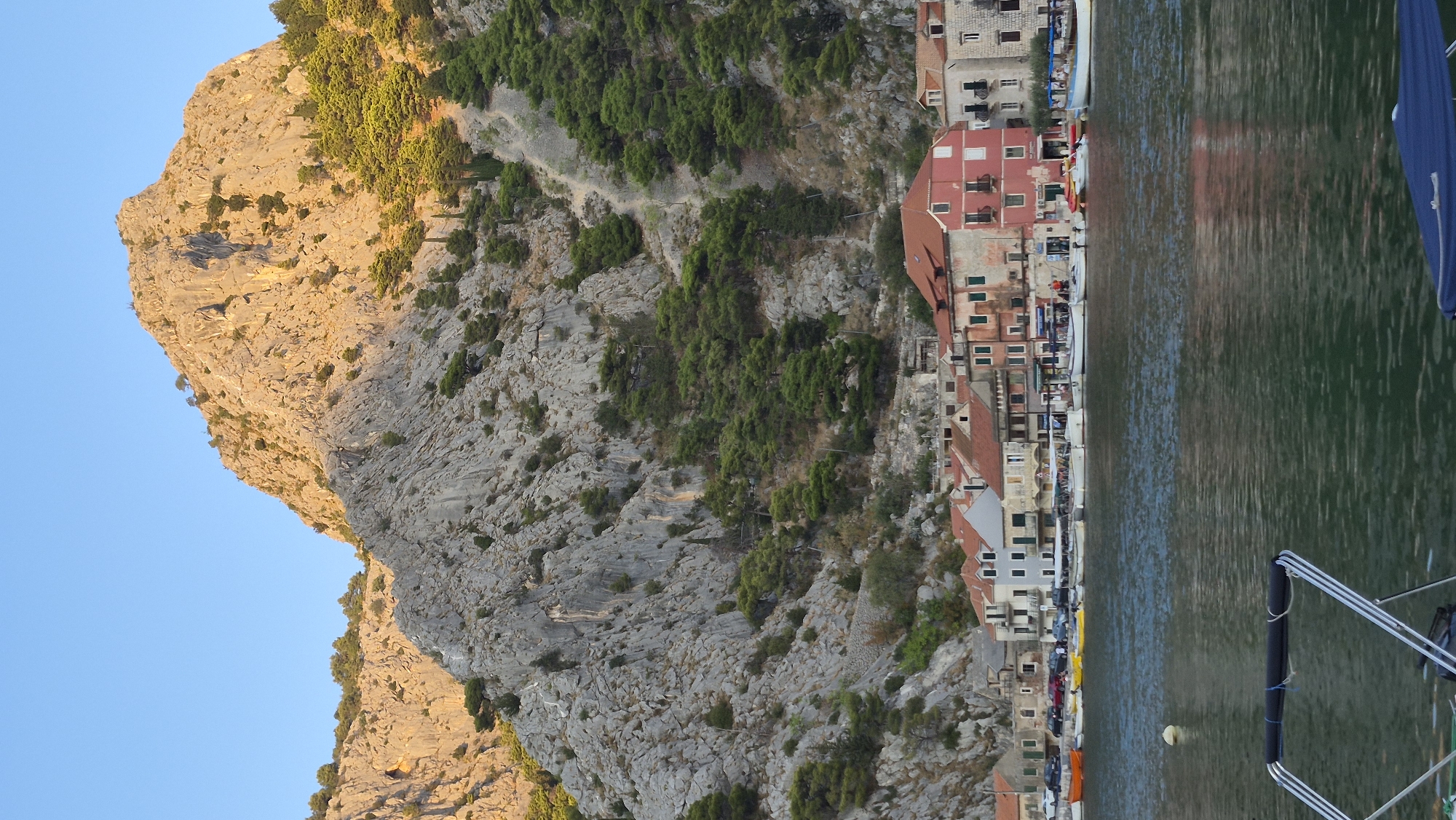 The image shows a bay with several boats docked along the waterfront of a town. The town has buildings with red tile roofs and a prominent pink building. Behind the town, a large, rocky mountain rises steeply, with sparse green trees growing on its slopes. The sky is clear and blue. The overall scene is a scenic view of a coastal town nestled against a mountainous backdrop.